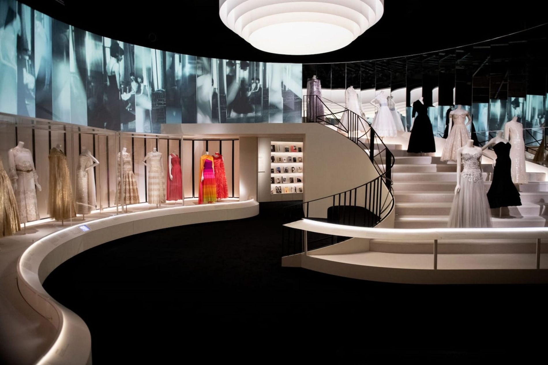 View of the final gallery, The Revived Allure, with many Chanel pieces on display around the room and up a sweeping staircase, there are projections from archive footage of fashion shows in Chanel's salon, these images are reflected in a mirrored wall facing the screens. The scene references Chanel's salon in Paris. From Gabrielle Chanel. Fashion Manifesto at the V&A Museum London. Soundscape by Coda to Coda.