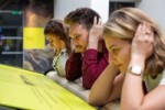 Image shows three people demonstrating how the bone conduction installation works - they are all leaning with their elbows on a soundbar and making a connection with their inner ear by using their hands. Installation Composer and Design by Coda to Coda