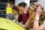 Image shows three people demonstrating how the bone conduction installation works - they are all leaning with their elbows on a soundbar and making a connection with their inner ear by using their hands. Installation Composer and Design by Coda to Coda