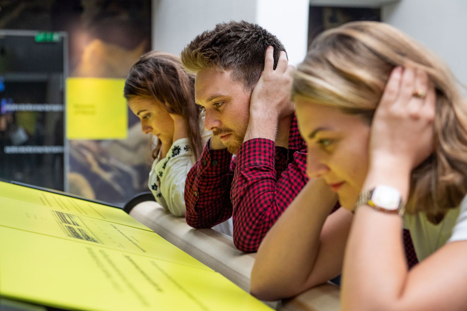 Image shows three people demonstrating how the bone conduction installation works - they are all leaning with their elbows on a soundbar and making a connection with their inner ear by using their hands. Installation Composer and Design by Coda to Coda
