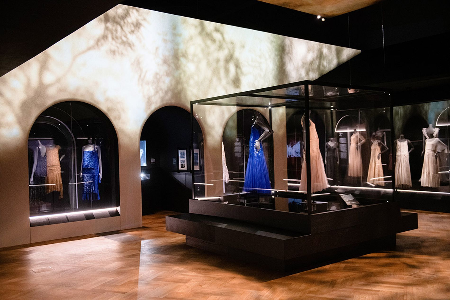 Image shows a view of the gallery, The Expression Of A Stark Luxury , with a blue and pink dress on display. The scene design reference's Chanel's house La Pausa. From Gabrielle Chanel. Fashion Manifesto at the V&A Museum London. Soundscape by Coda to Coda.