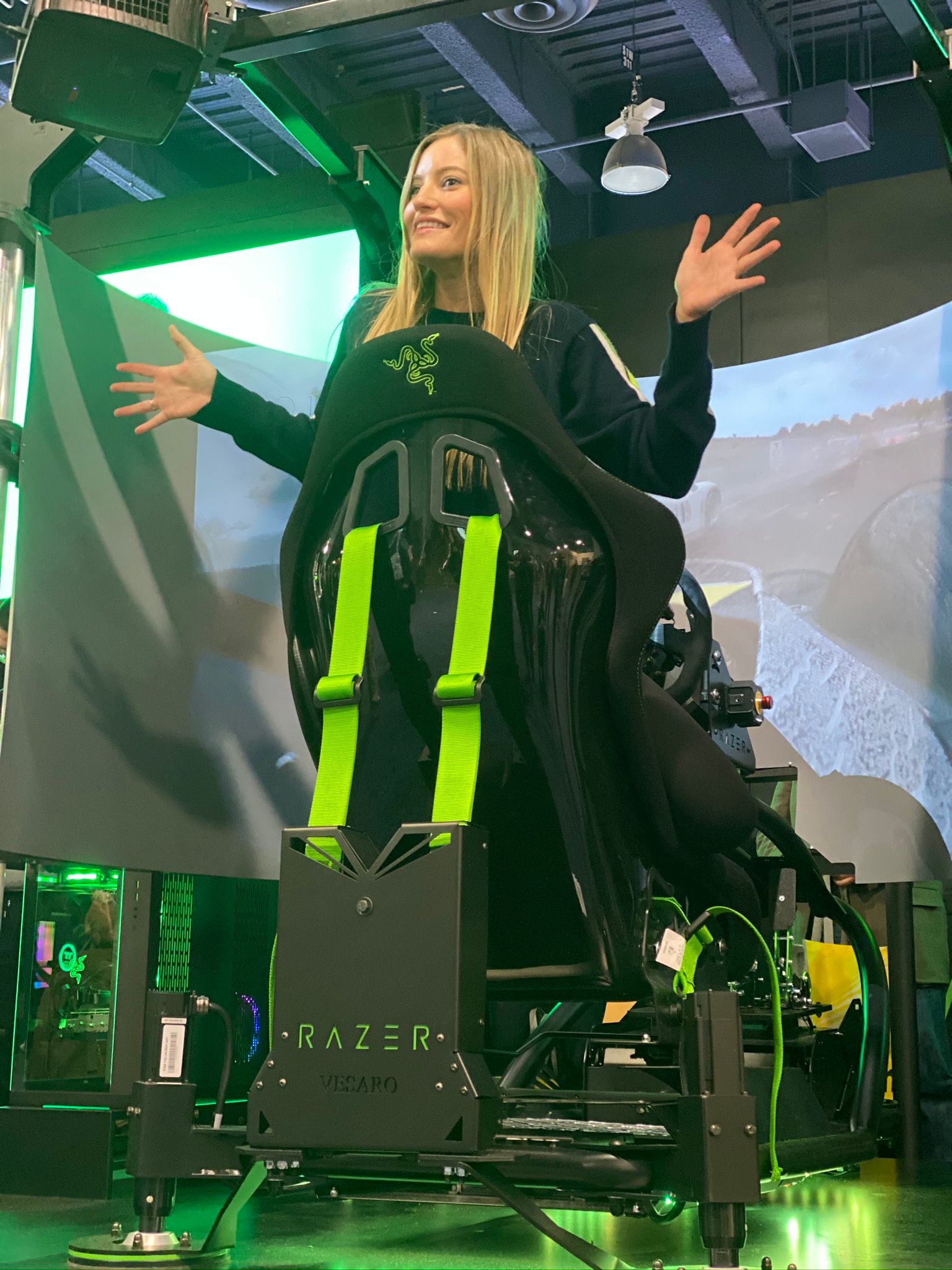 Woman with arms out seated in a Razer-branded motion simulator seat with green harness