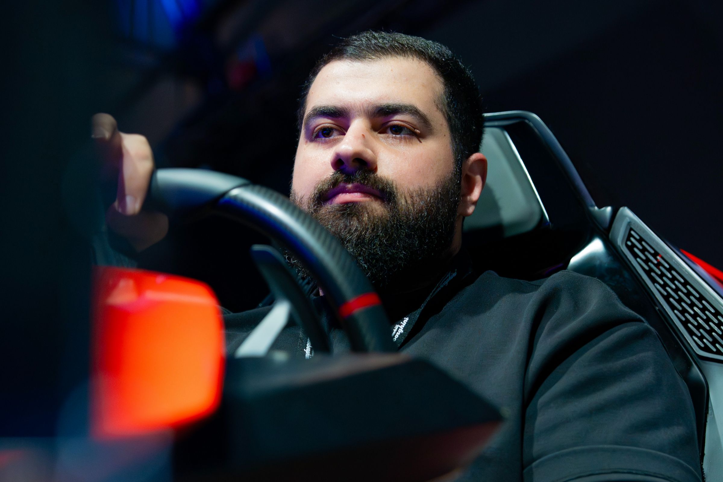 Bearded man in a black top at the wheel of a Lamborghini-styled simulator under stage lighting