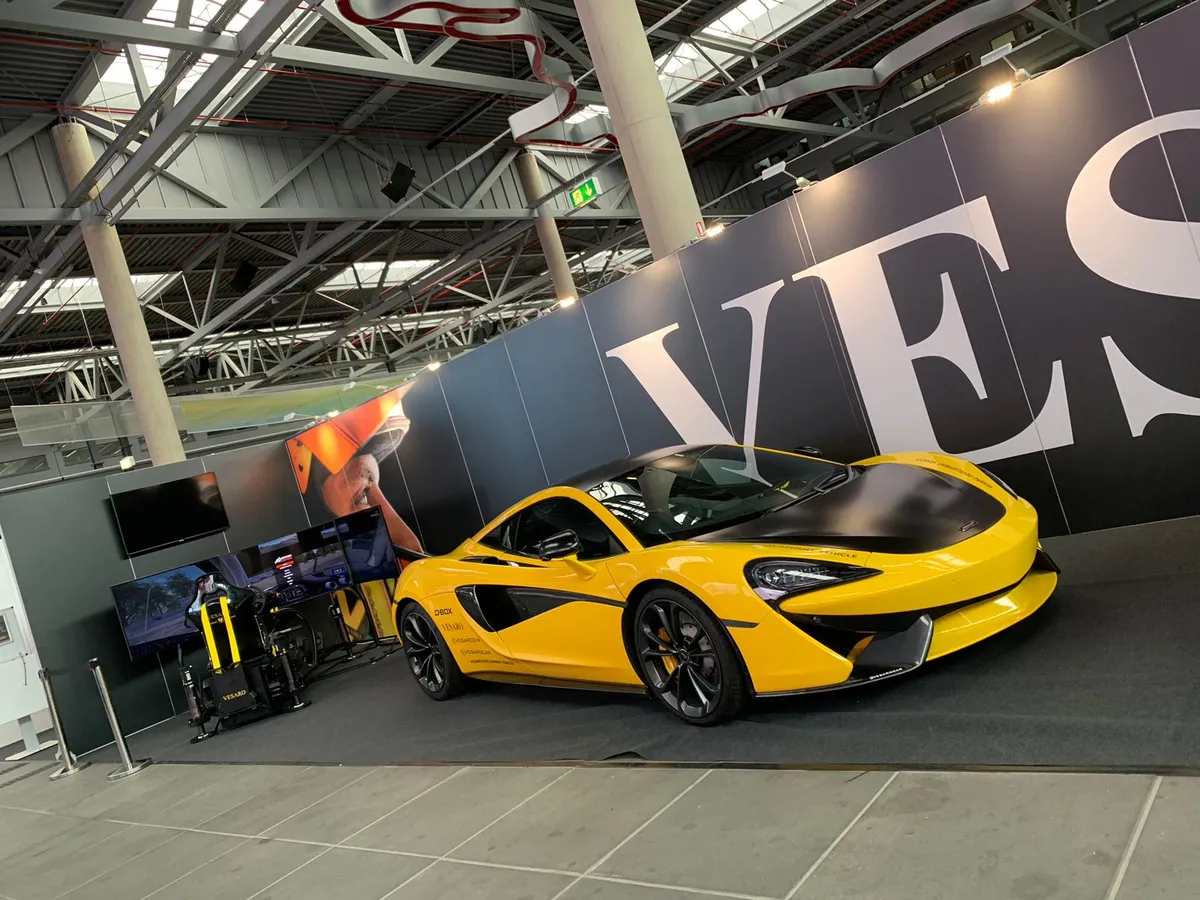 Yellow McLaren sports car parked on an exhibition stand beside a simulator rig and a large banner