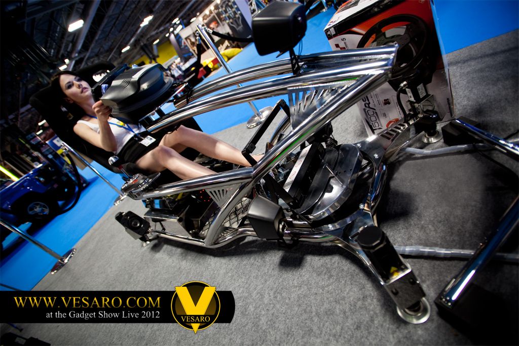 Woman seated in a chrome-framed Vesaro simulator rig at the Gadget Show Live 2012 stand, seen from a low angle