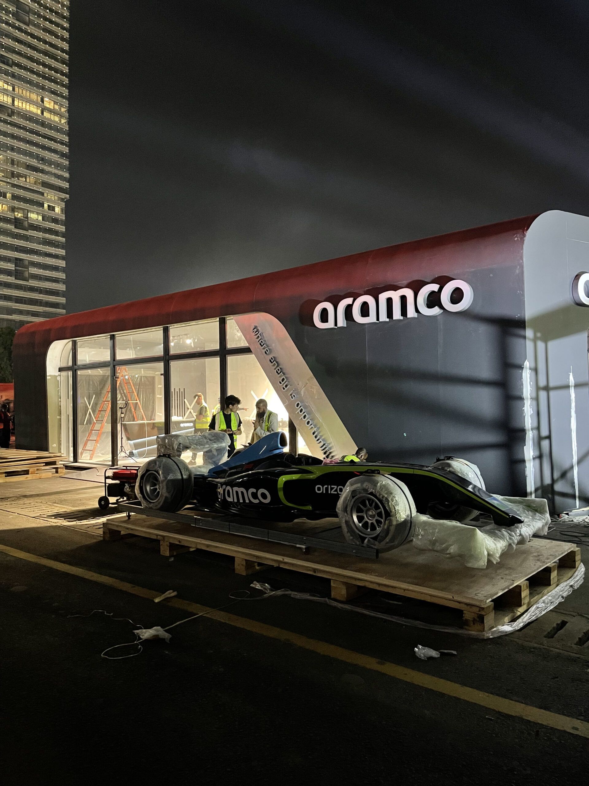 Aramco-branded racing car on a wooden pallet outside an Aramco pavilion at night