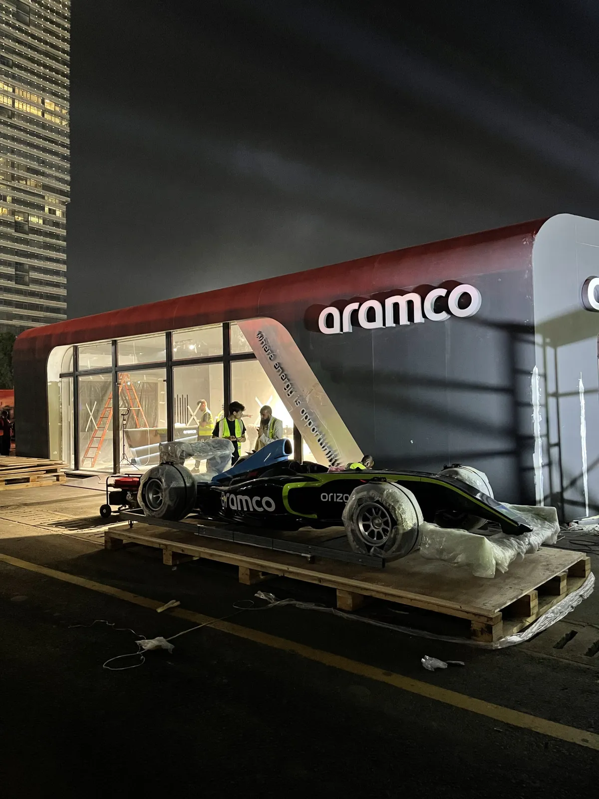 Aramco-branded racing car on a wooden pallet outside an Aramco pavilion at night
