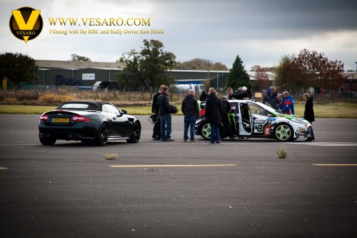 Crew and onlookers on an airfield beside a Jaguar and a rally car during BBC filming with Ken Block