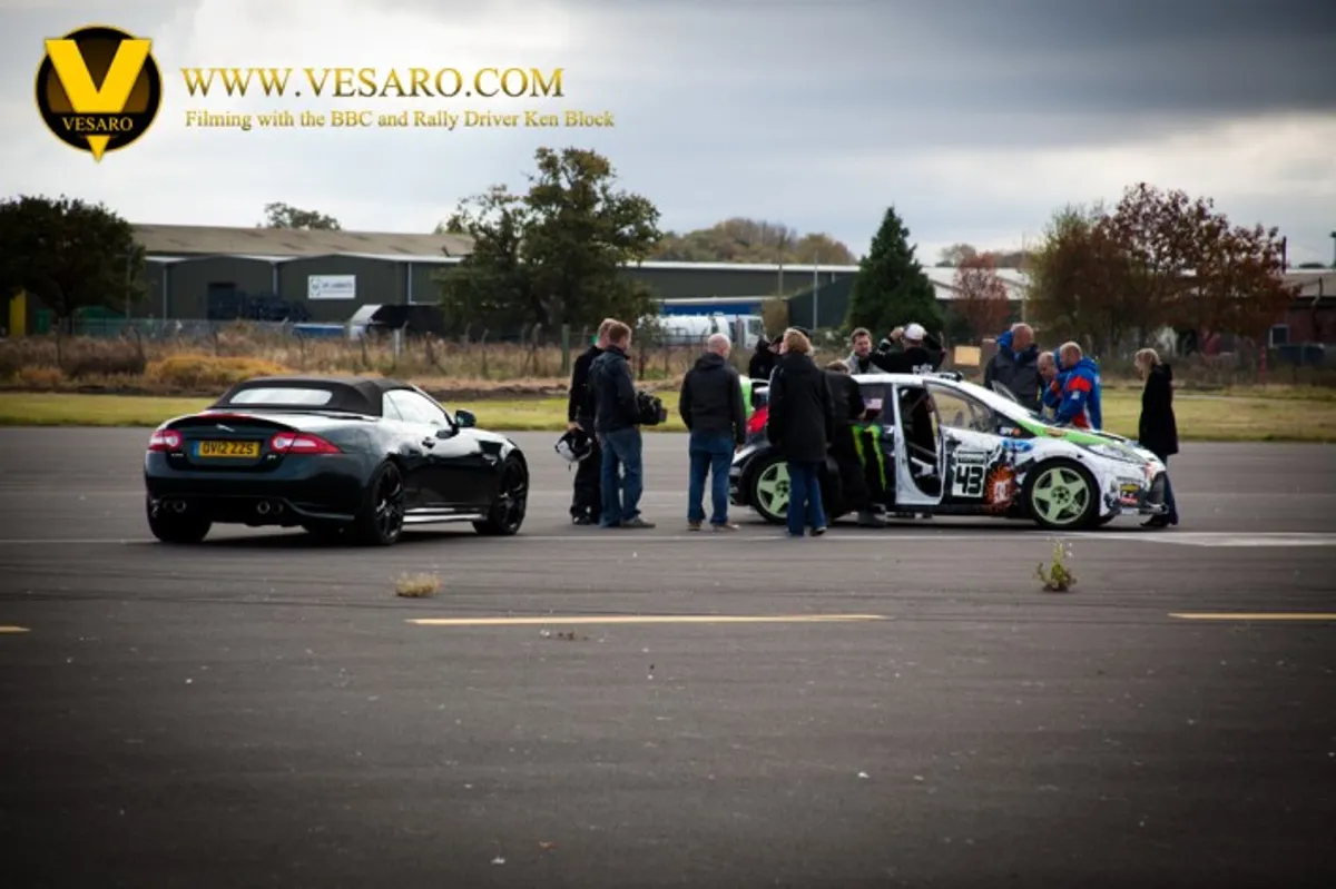 Crew and onlookers on an airfield beside a Jaguar and a rally car during BBC filming with Ken Block