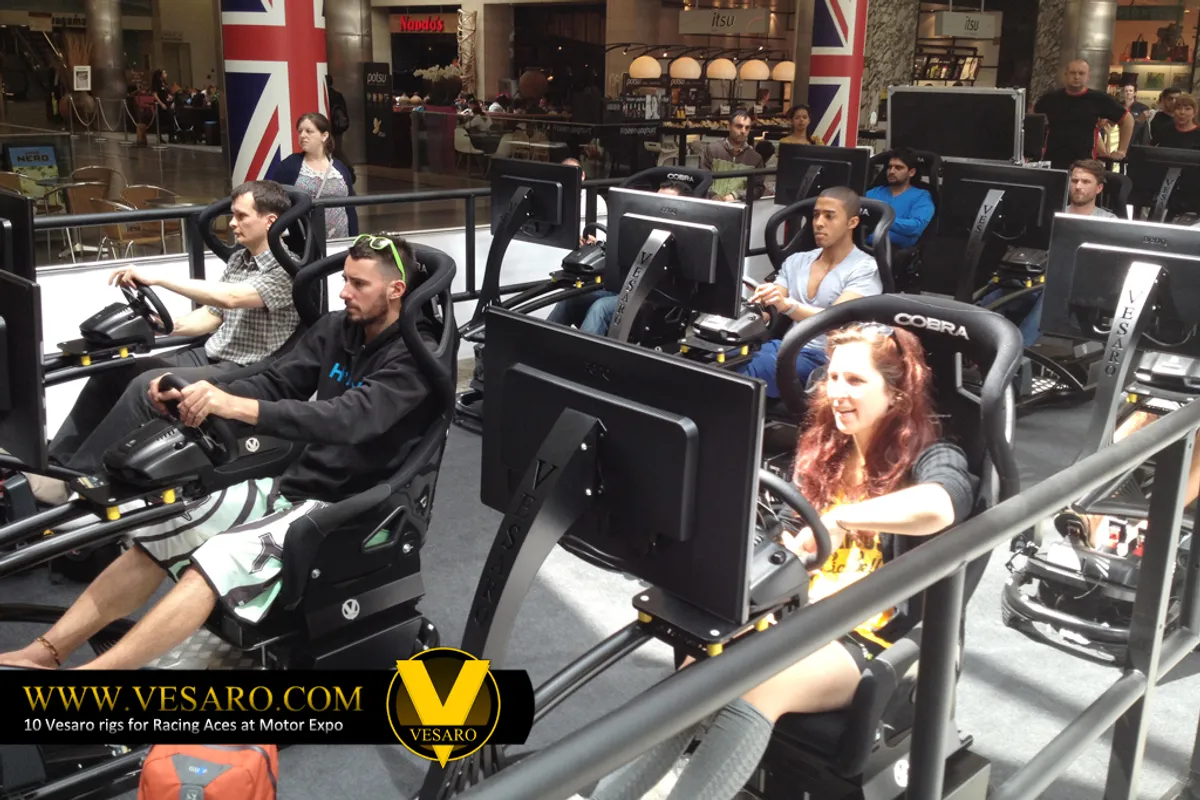 Row of people seated at Vesaro racing simulators with monitors on a shopping centre floor, Union Jack flags behind