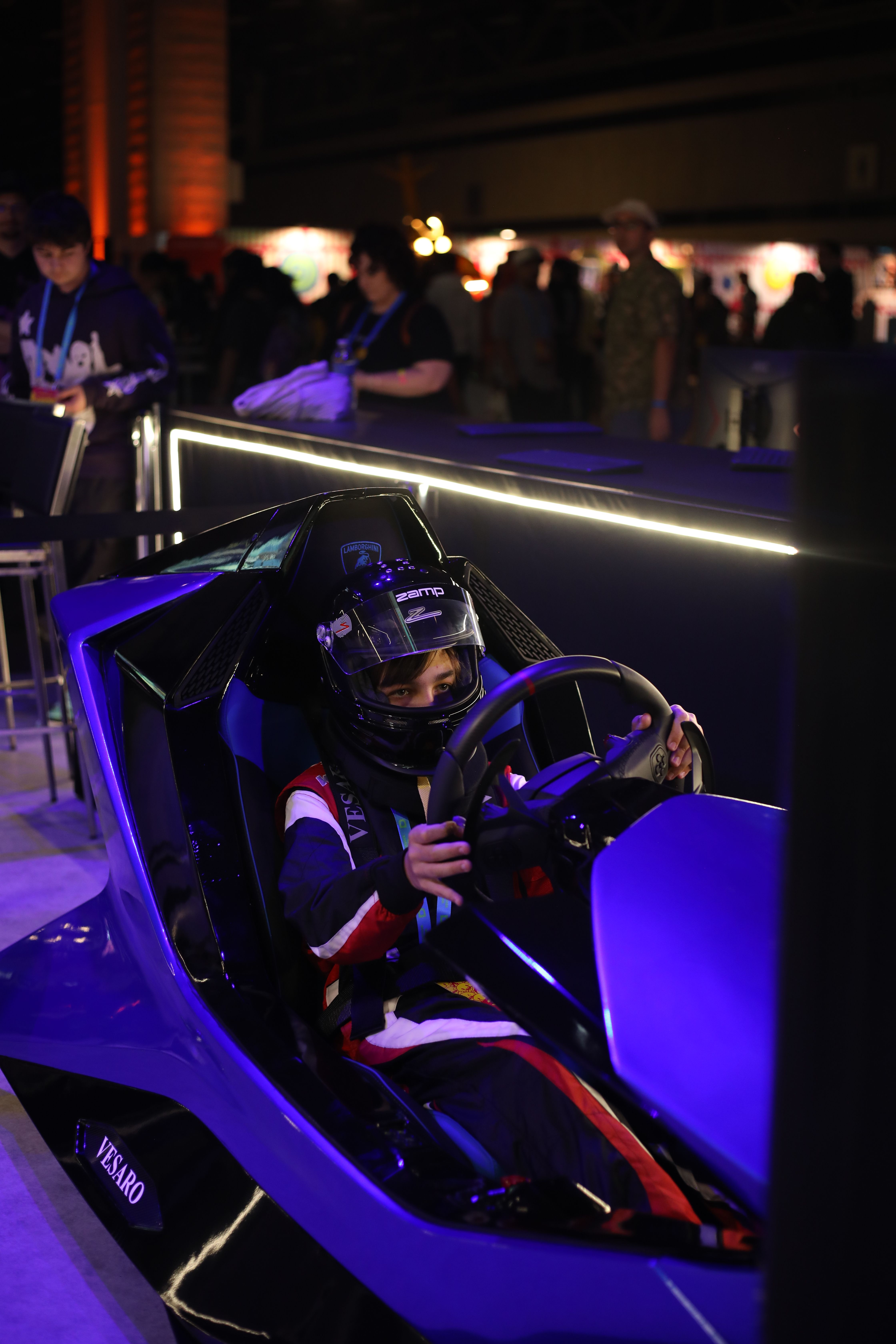 Driver in a racing helmet and race suit at the wheel of a blue Vesaro simulator under purple event lighting