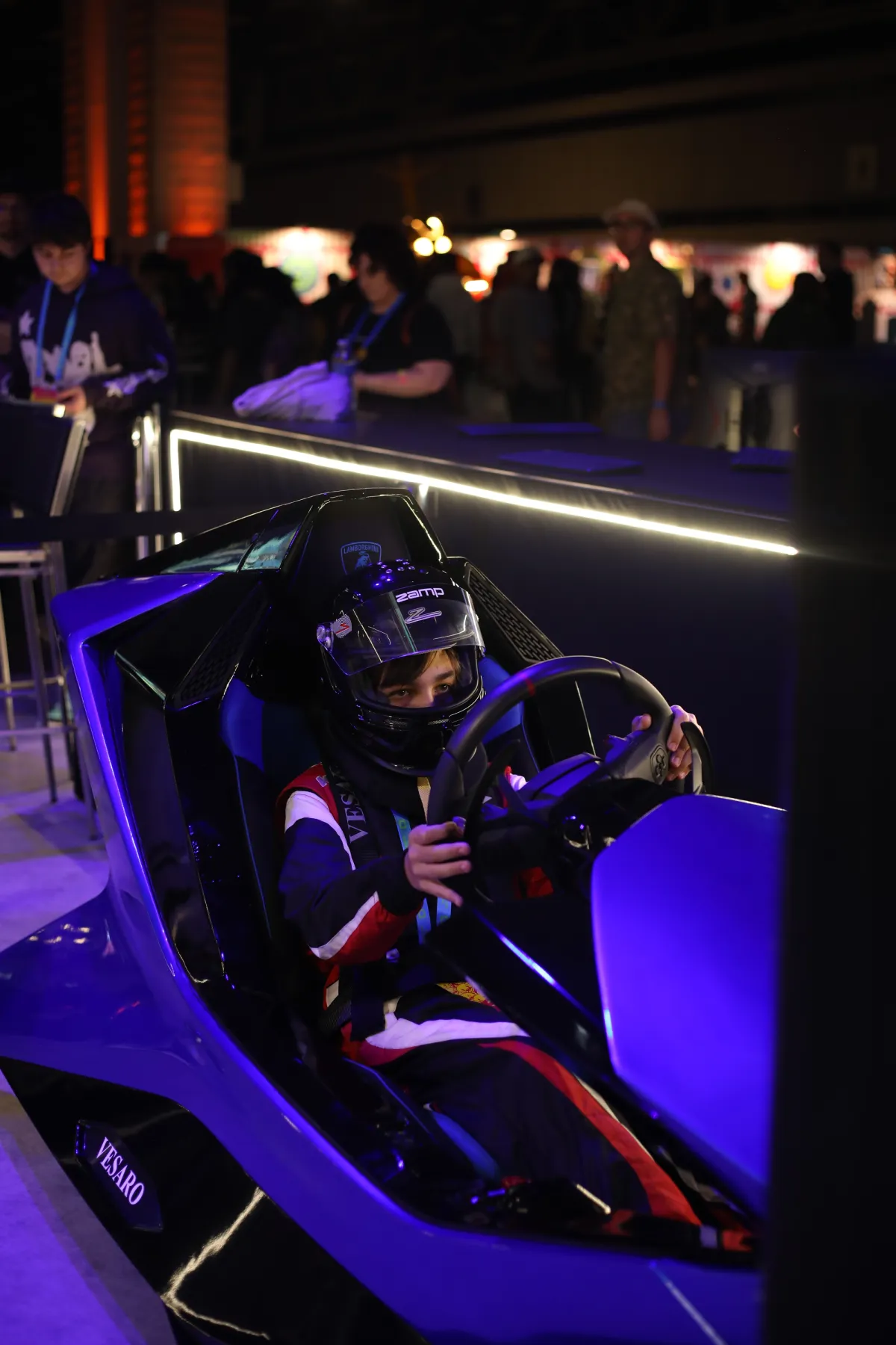Driver in a racing helmet and race suit at the wheel of a blue Vesaro simulator under purple event lighting