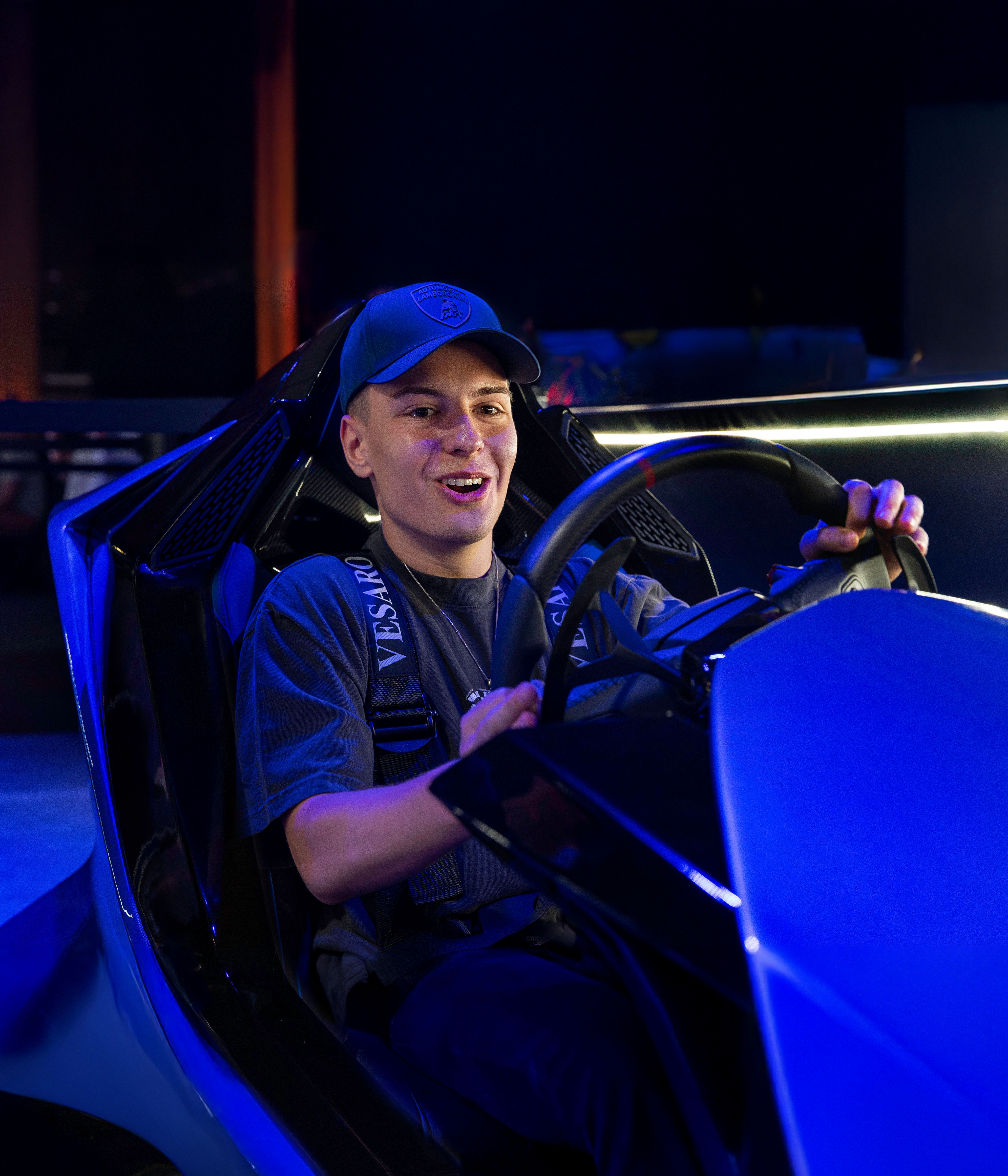 Smiling young man in a cap gripping the wheel of a blue racing simulator in dark blue lighting