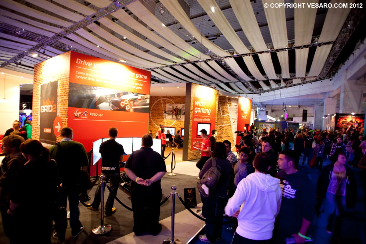 Busy exhibition stand with red and orange display panels and crowds queuing at a racing game event