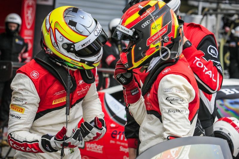 Two racing drivers in colourful helmets and red race suits talking in the pit lane