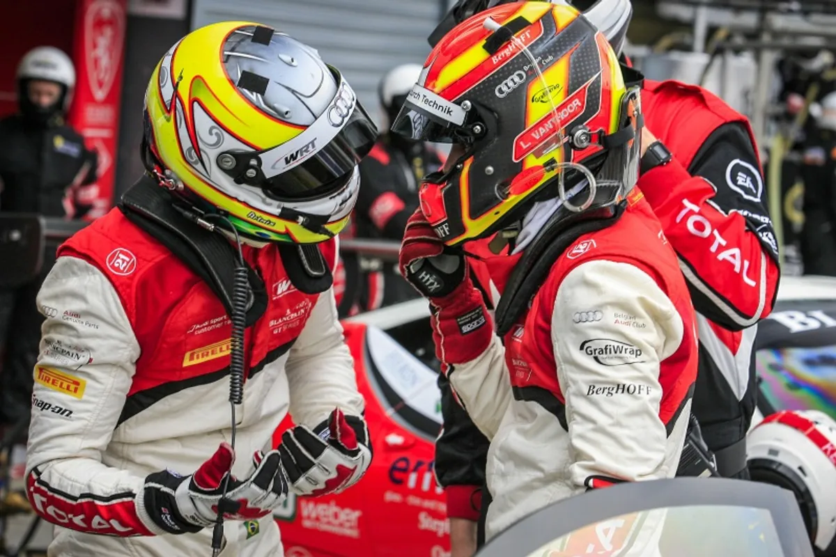 Two racing drivers in colourful helmets and red race suits talking in the pit lane
