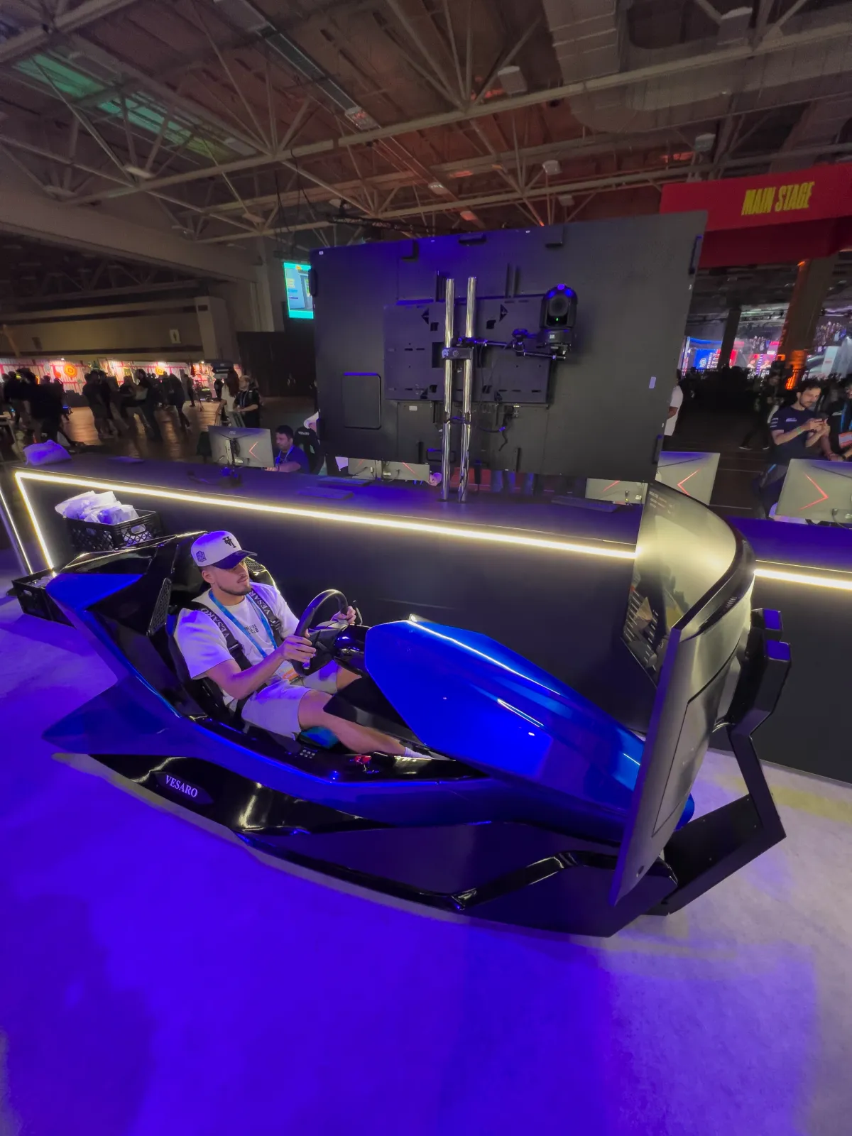 Person driving a blue Vesaro simulator with a display behind it, purple lit exhibition hall in the background