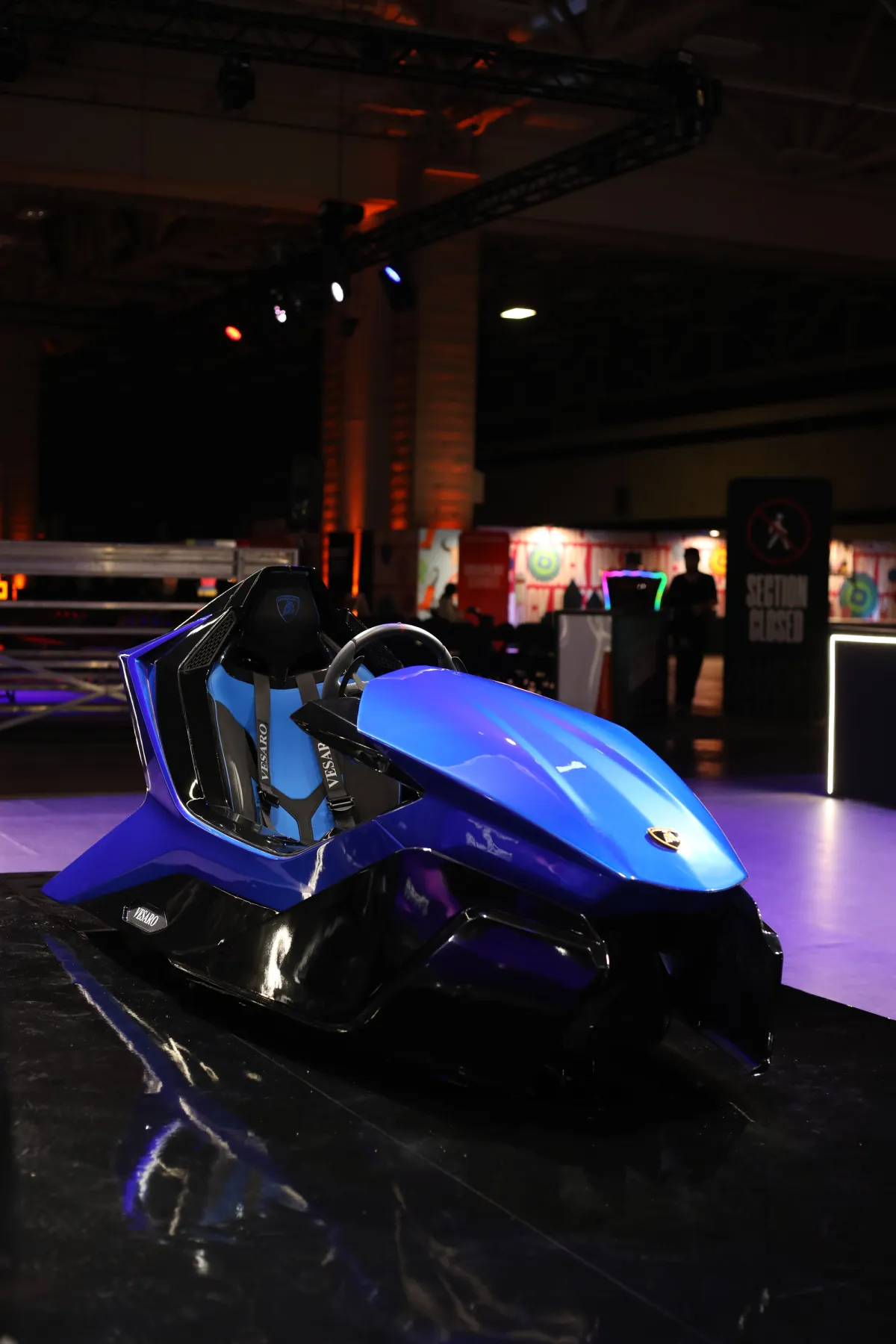 Empty blue Vesaro x Lamborghini simulator on a glossy black plinth in a dark exhibition hall