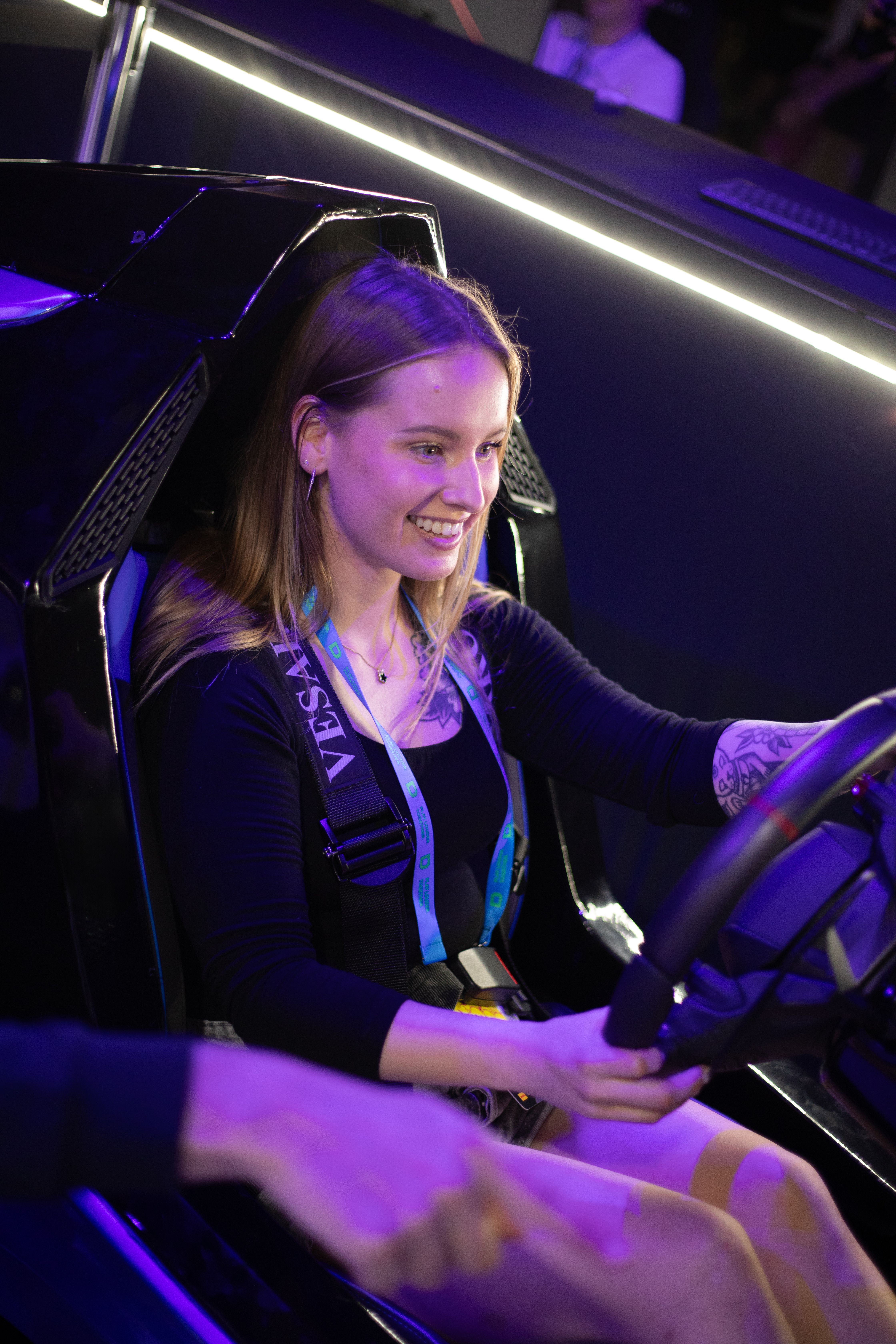 Smiling woman in a lanyard seated in a black racing simulator seat, hands on the wheel