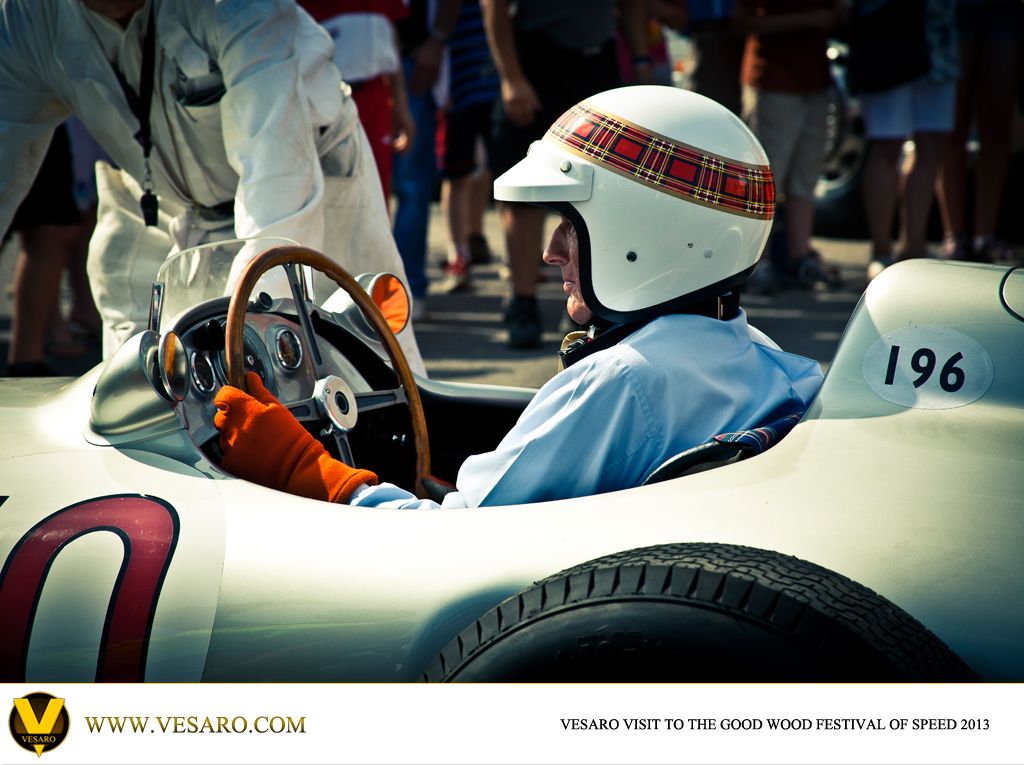 Driver in a tartan-banded helmet and wooden steering wheel seated in a vintage Mercedes racing car