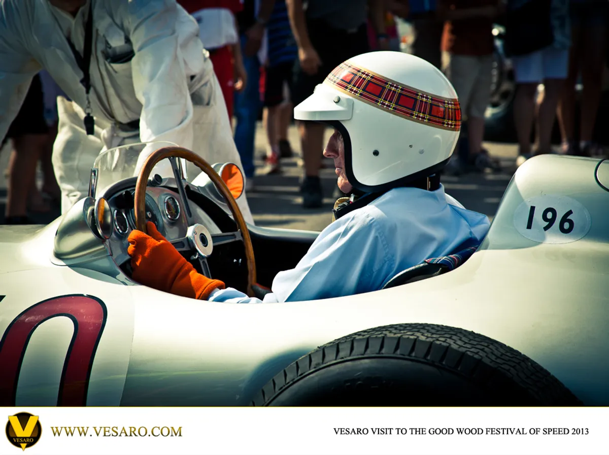 Driver in a tartan-banded helmet and wooden steering wheel seated in a vintage Mercedes racing car