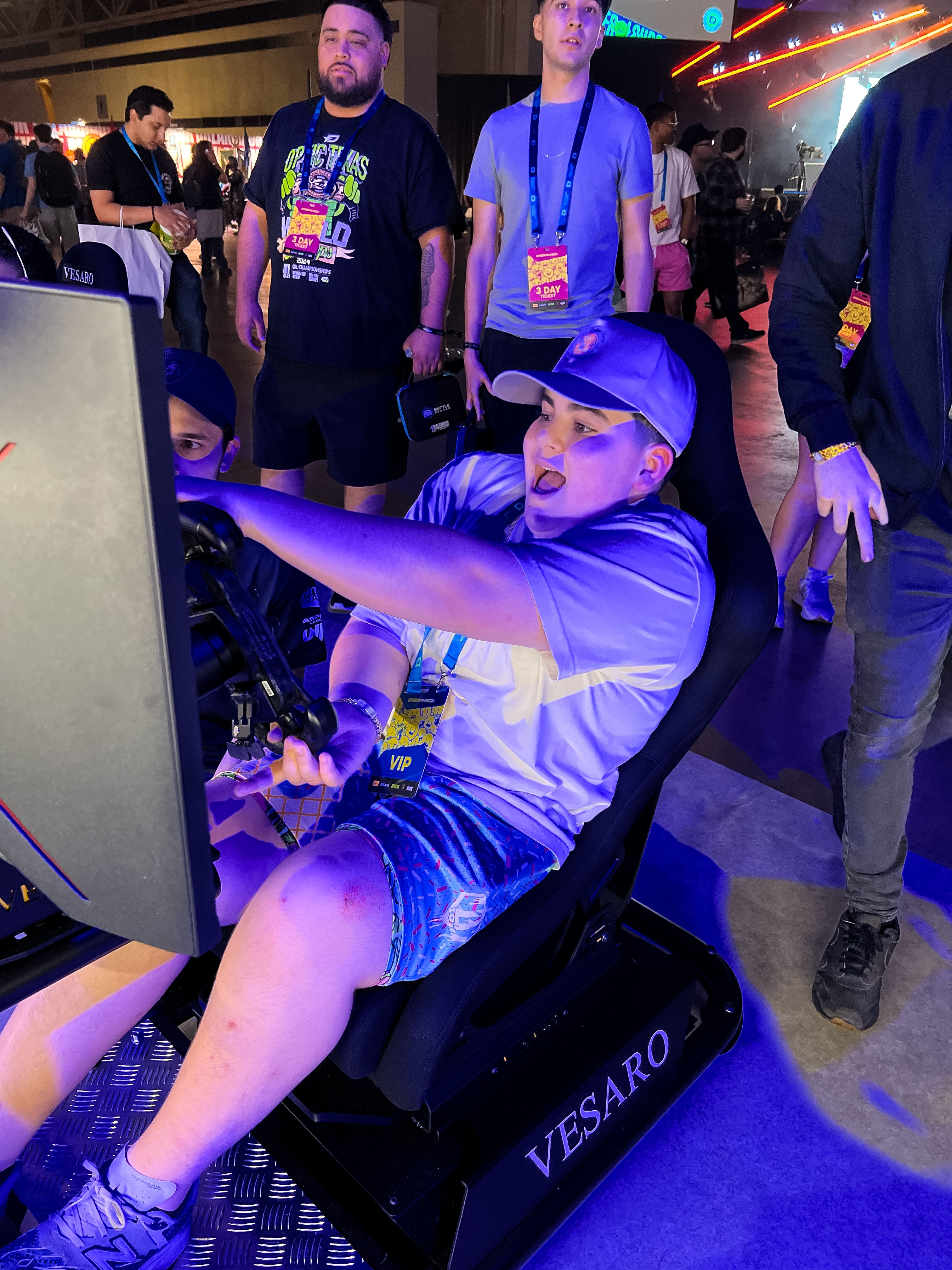 Young man in a cap driving a Vesaro simulator with a Vesaro-branded seat base at a busy gaming event