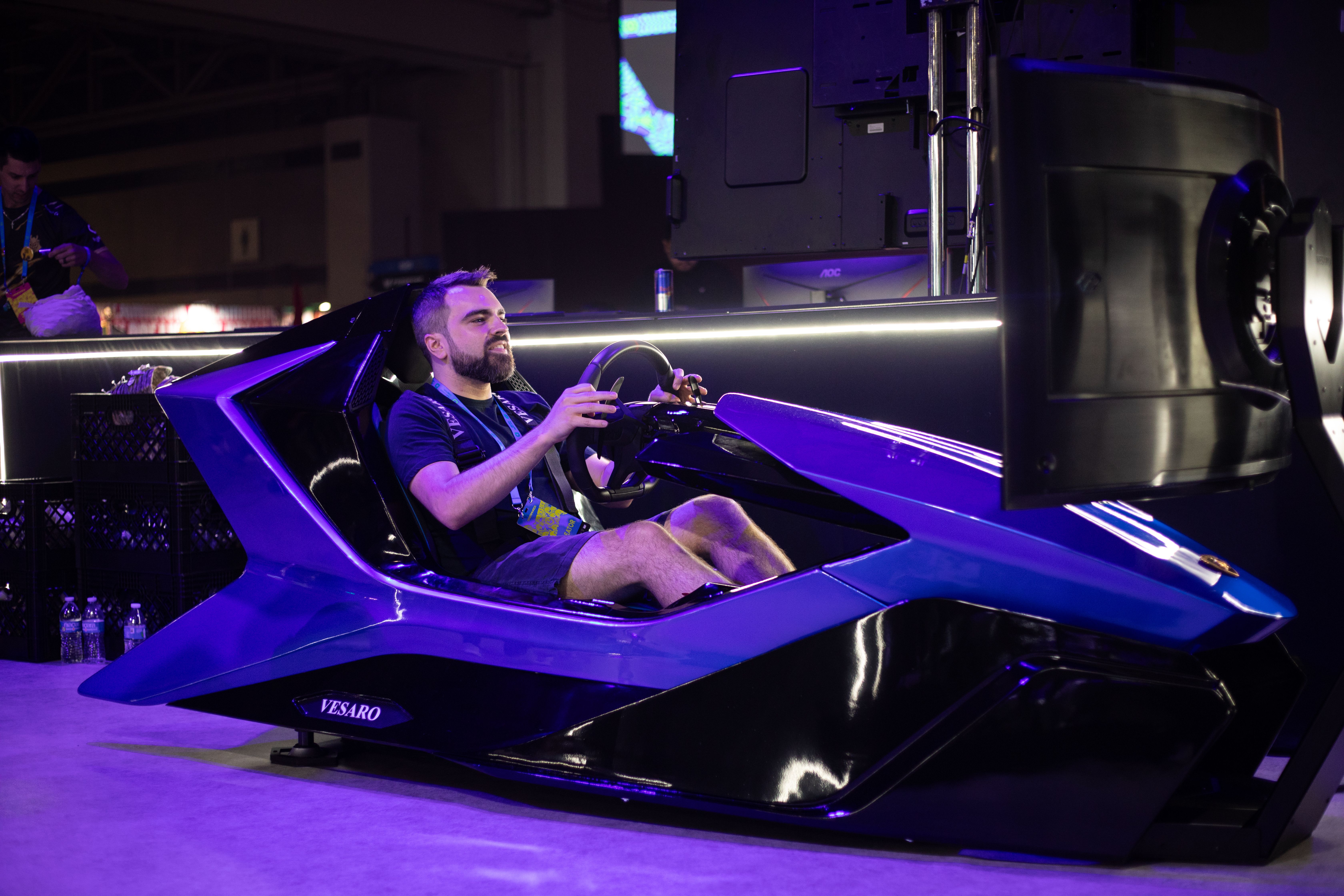 Bearded man in a lanyard at the wheel of a purple and black Vesaro simulator beside a display