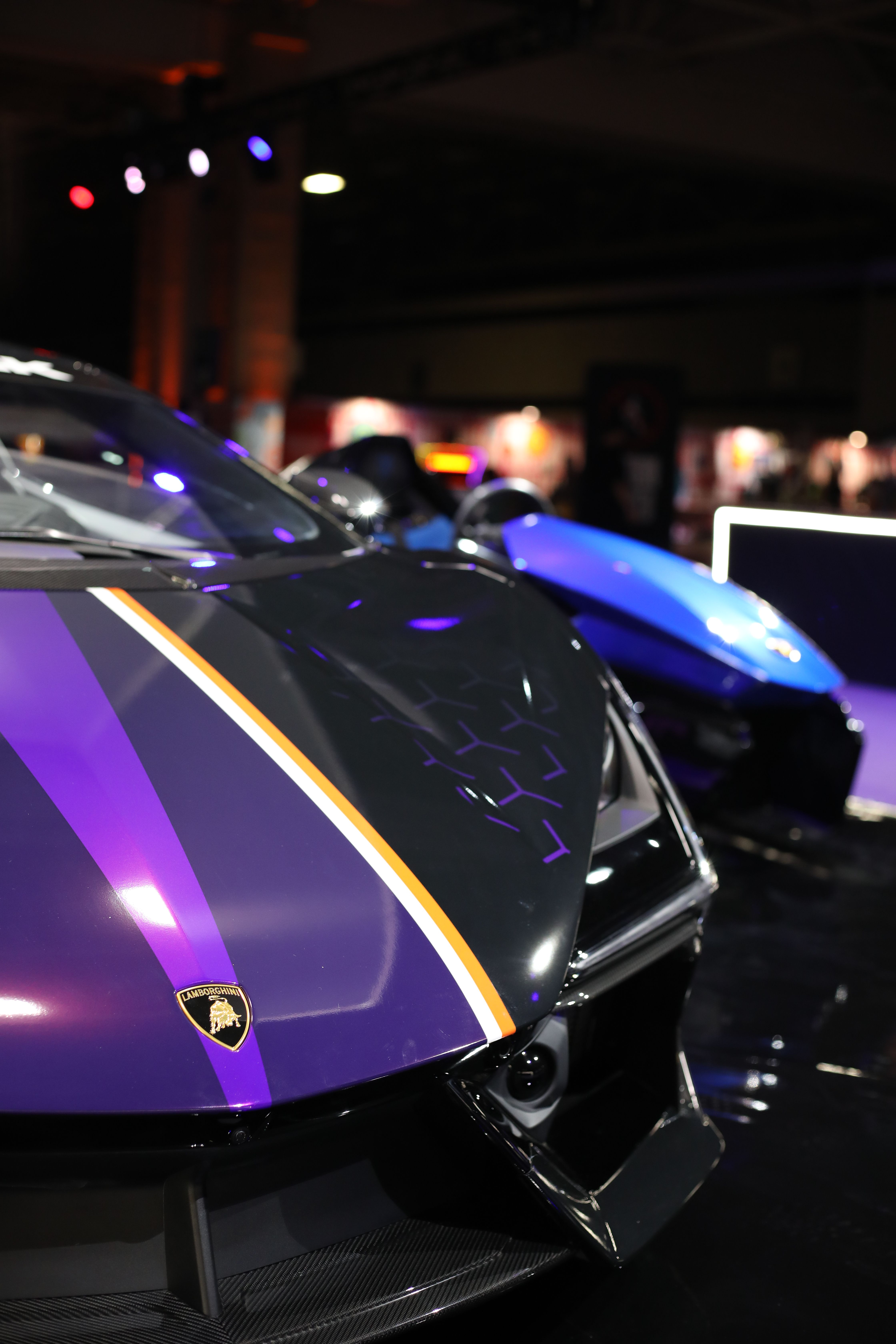 Front of a black and purple Lamborghini supercar with gold stripe and shield badge, with a blue simulator behind