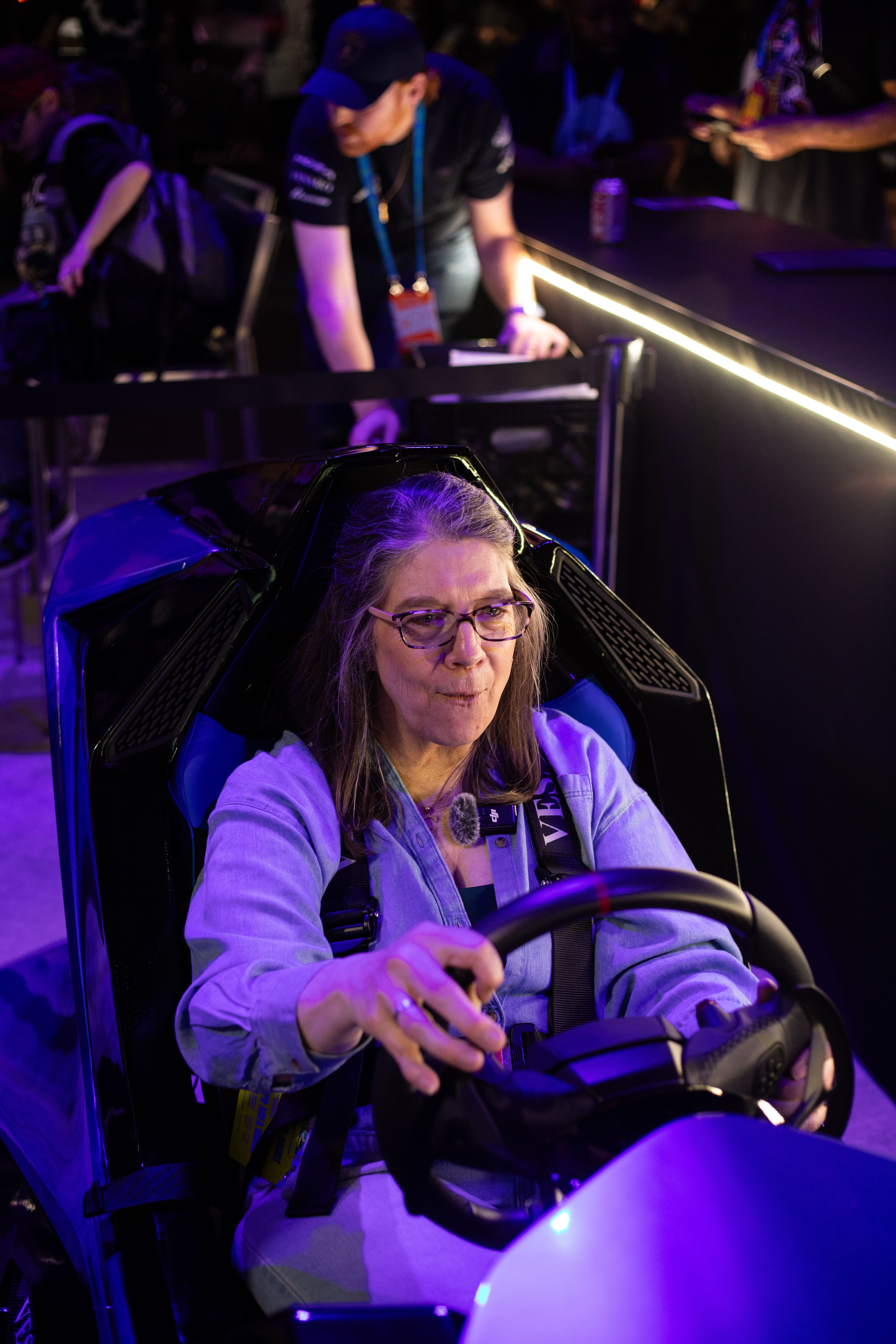 Woman with glasses gripping a steering wheel in a racing simulator seat under purple lighting at an event