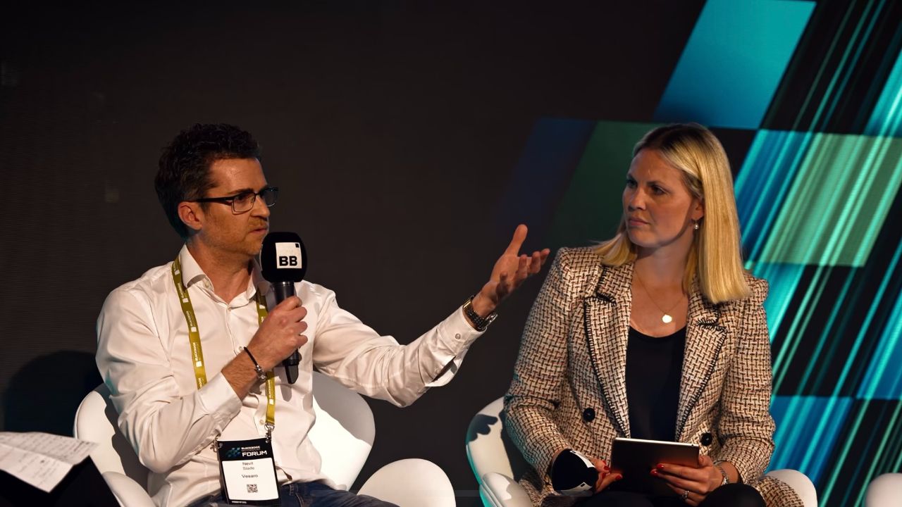 Man with a microphone and a woman with a notebook seated on stage during a panel discussion