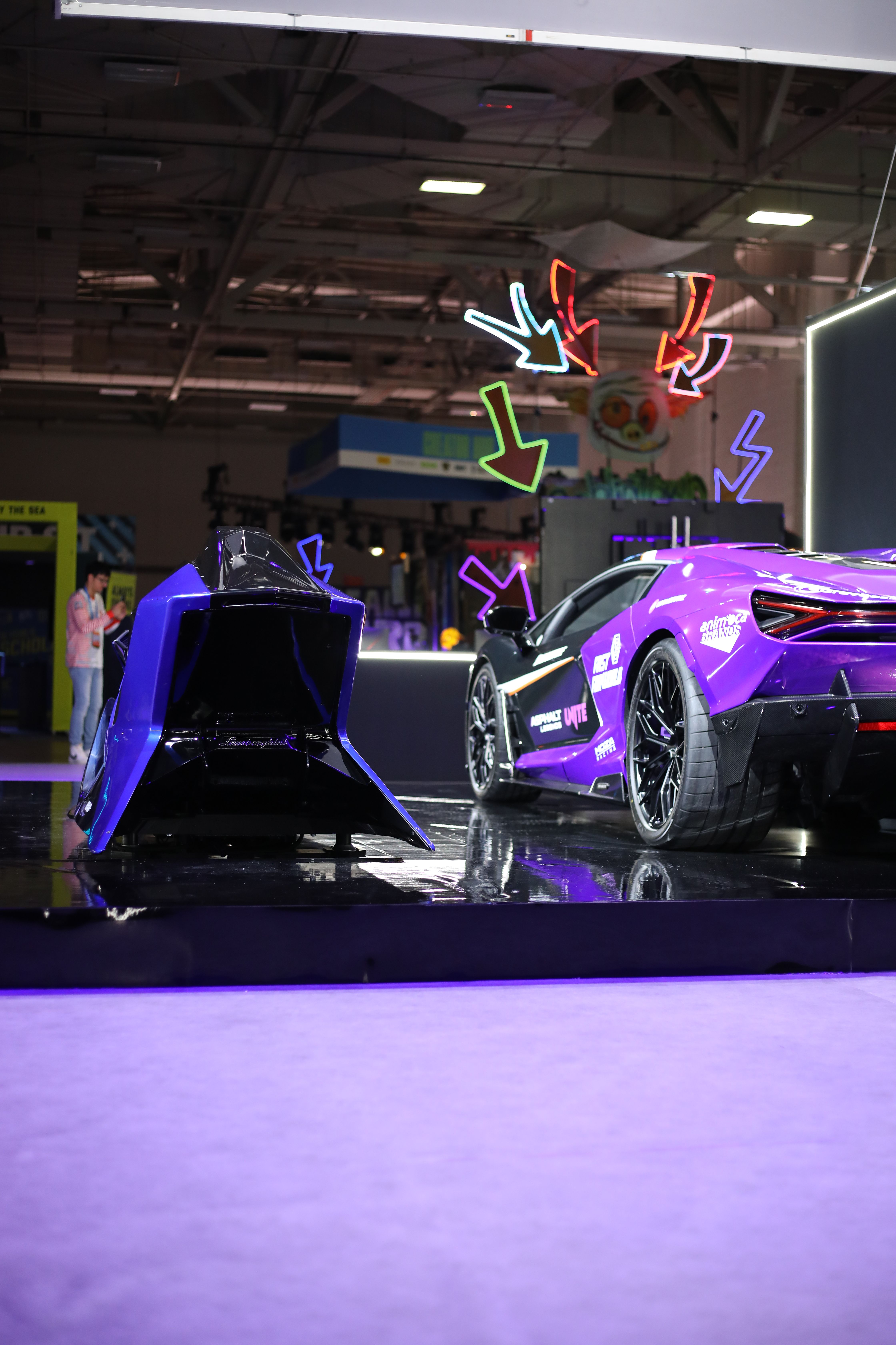 Purple Lamborghini supercar next to a blue simulator on a black plinth with neon arrow signs behind
