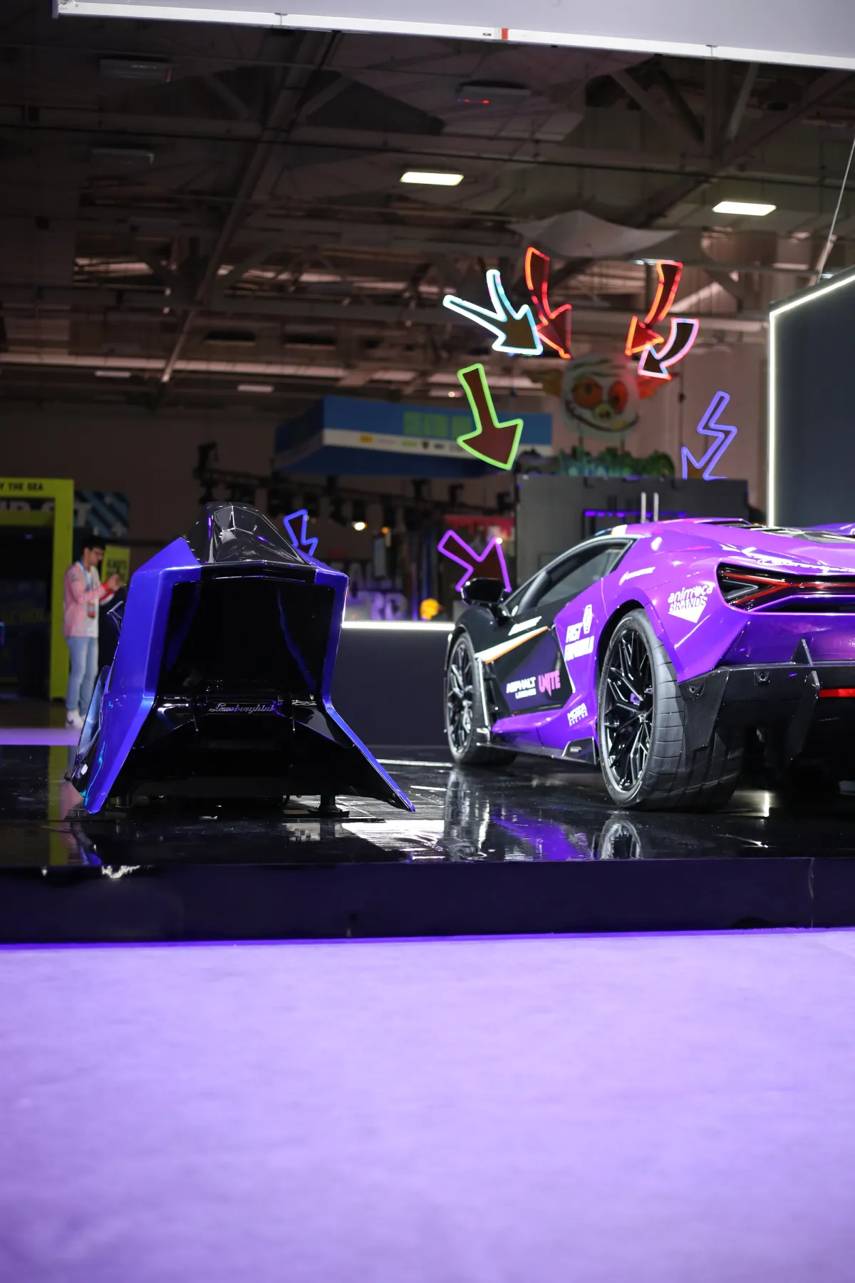 Purple Lamborghini supercar next to a blue simulator on a black plinth with neon arrow signs behind