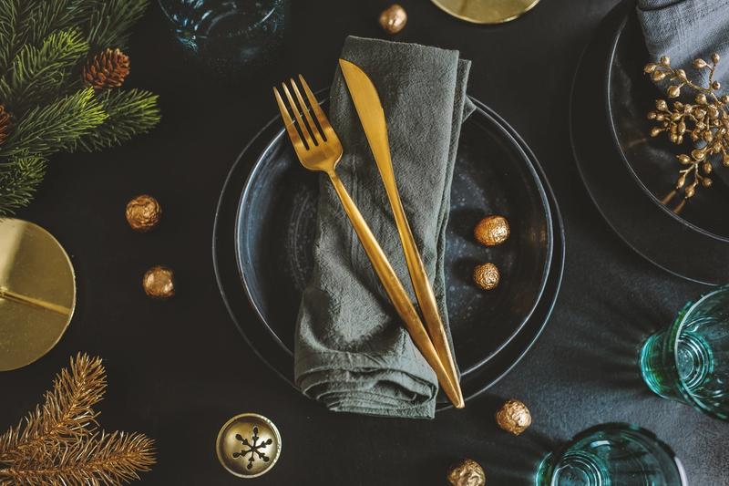 Cubiertos dorados sobre servilleta de tela y plato oscuro sobre mesa con decoración navideña