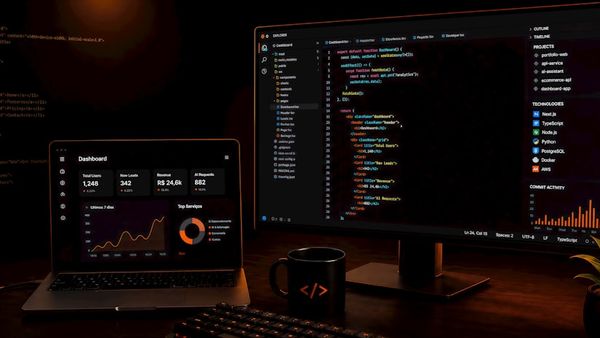 Dark workspace with laptop, monitor displaying code, and plant