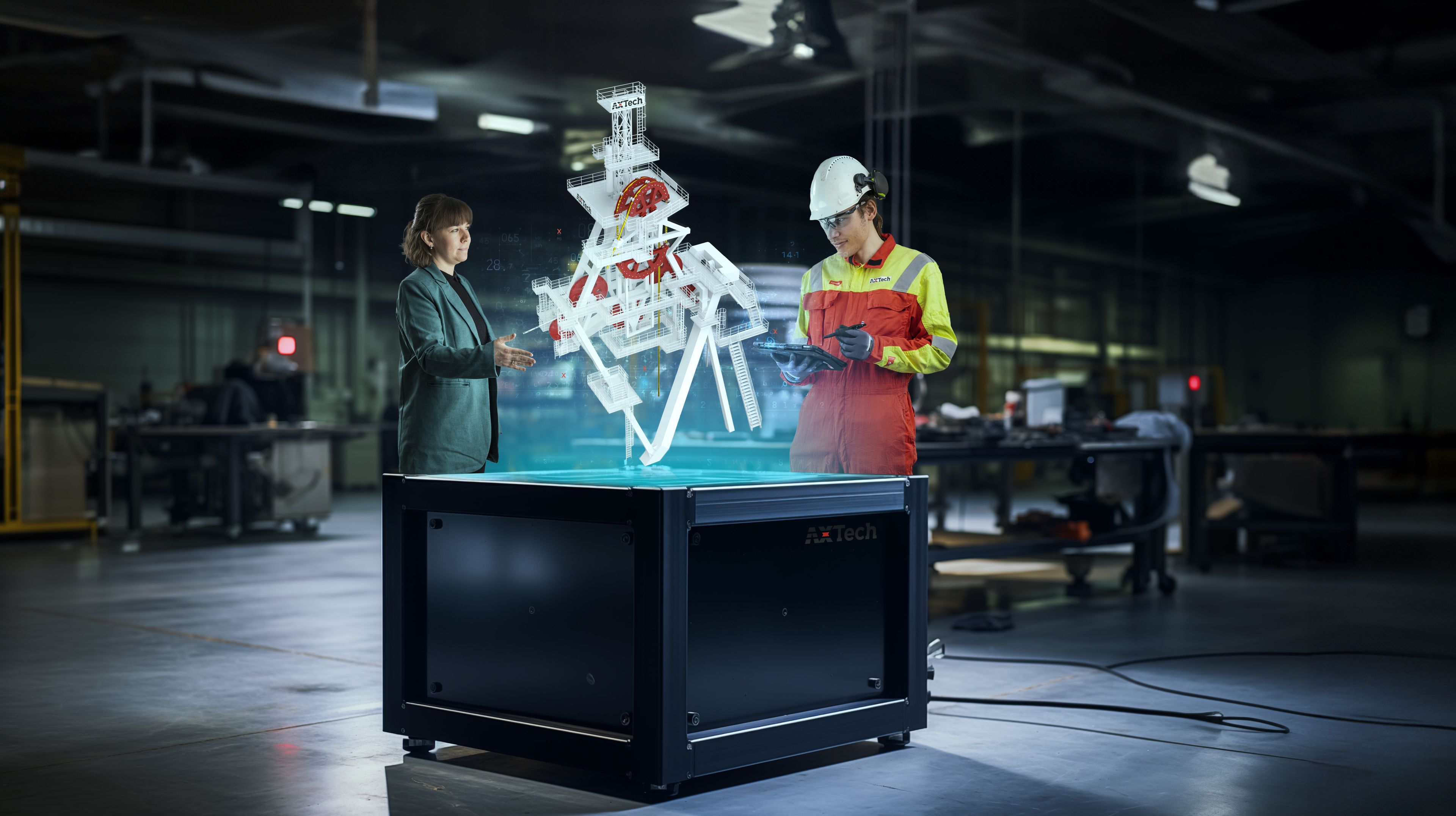 Two professionals interacting with a 3D holographic industrial rig model projected from a black rectangular device in a modern industrial workspace.