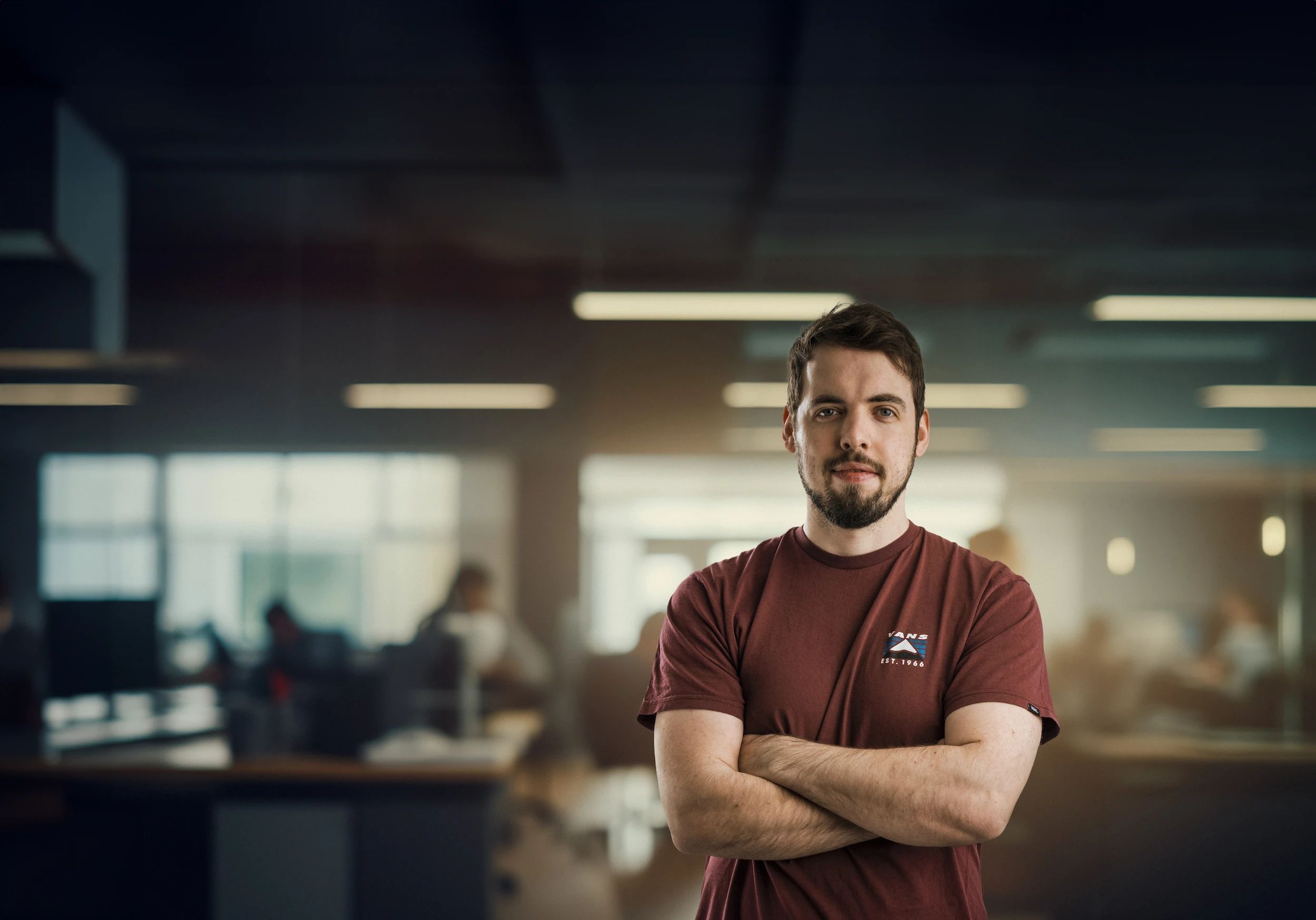 Young man with short dark hair and beard wearing a maroon Vans t-shirt stands with arms crossed in a modern office setting.