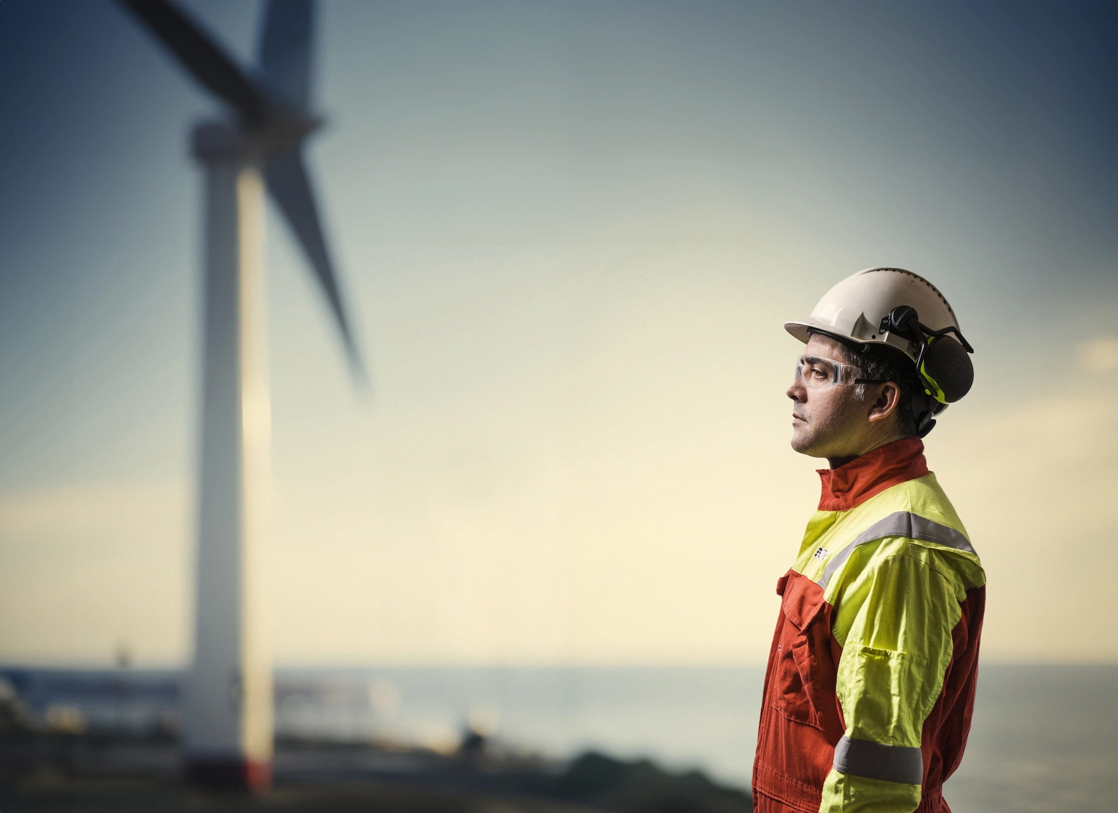 Engineer wearing safety gear and helmet looking at an offshore wind turbine at sunset.