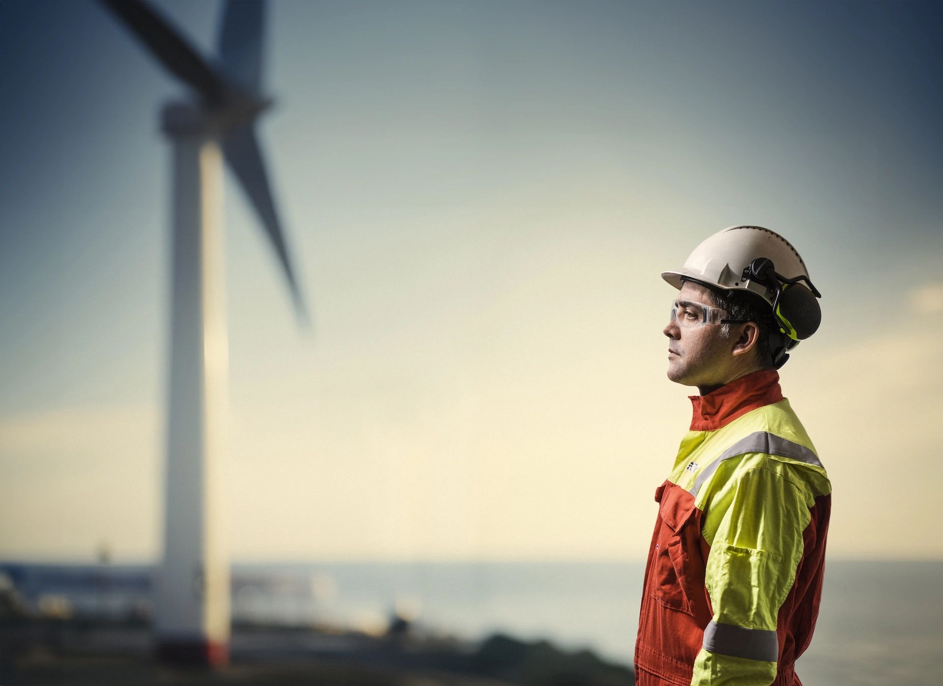 Engineer wearing safety gear and helmet looking at an offshore wind turbine at sunset.