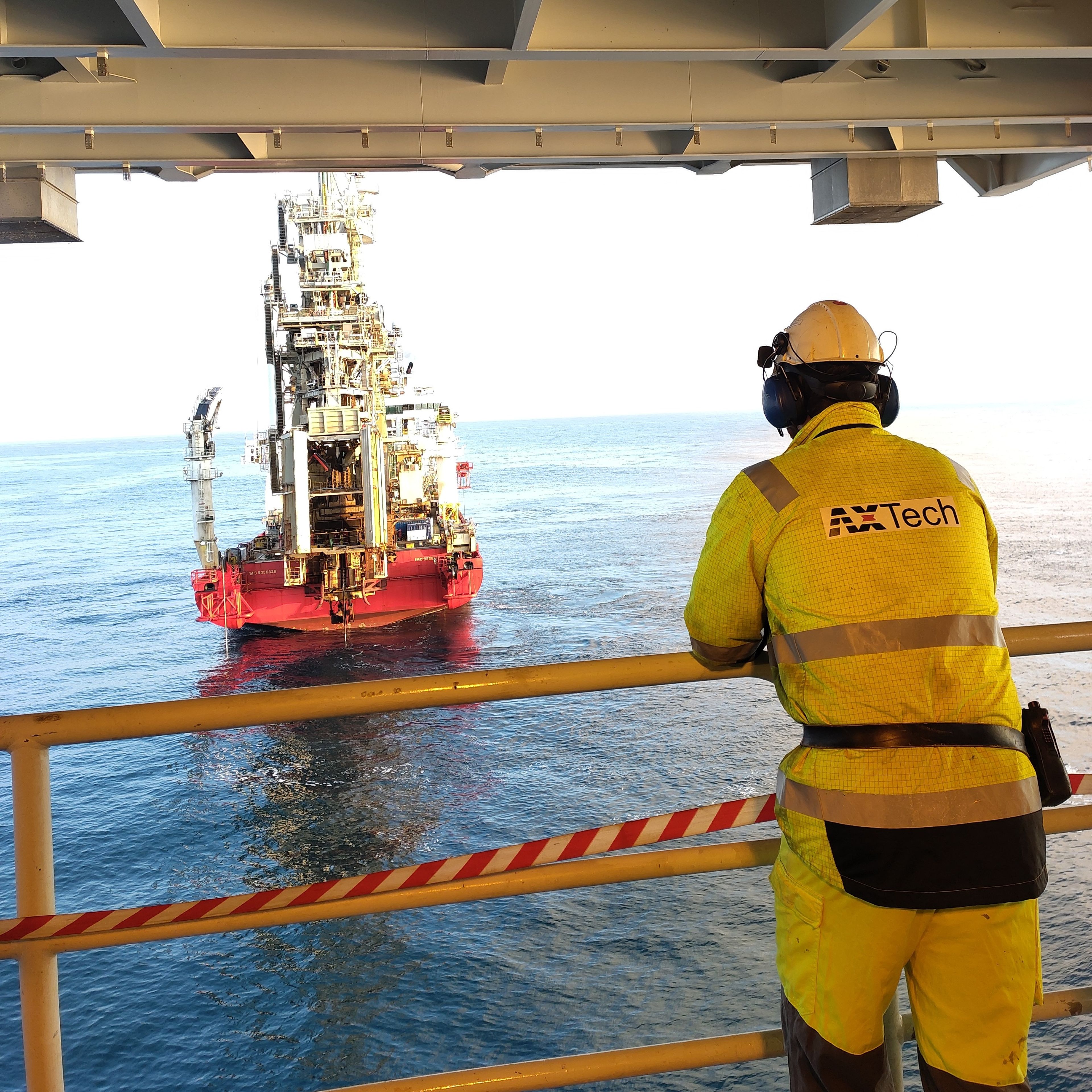 AXTech engineer in yellow coveralls and hard hat watching a subsea module handling operation at sea from an offshore platform.