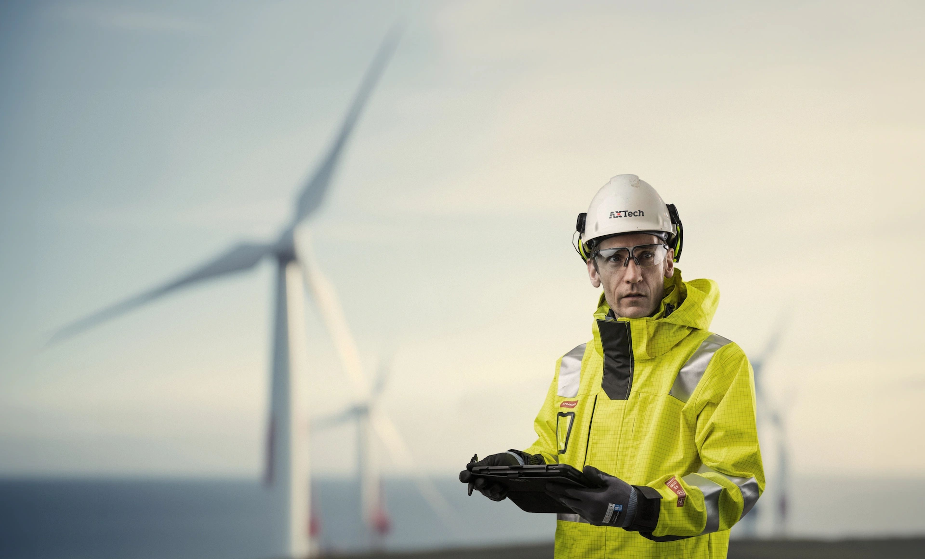 Engineer in high-visibility jacket and helmet holding tablet with wind turbines in the background.