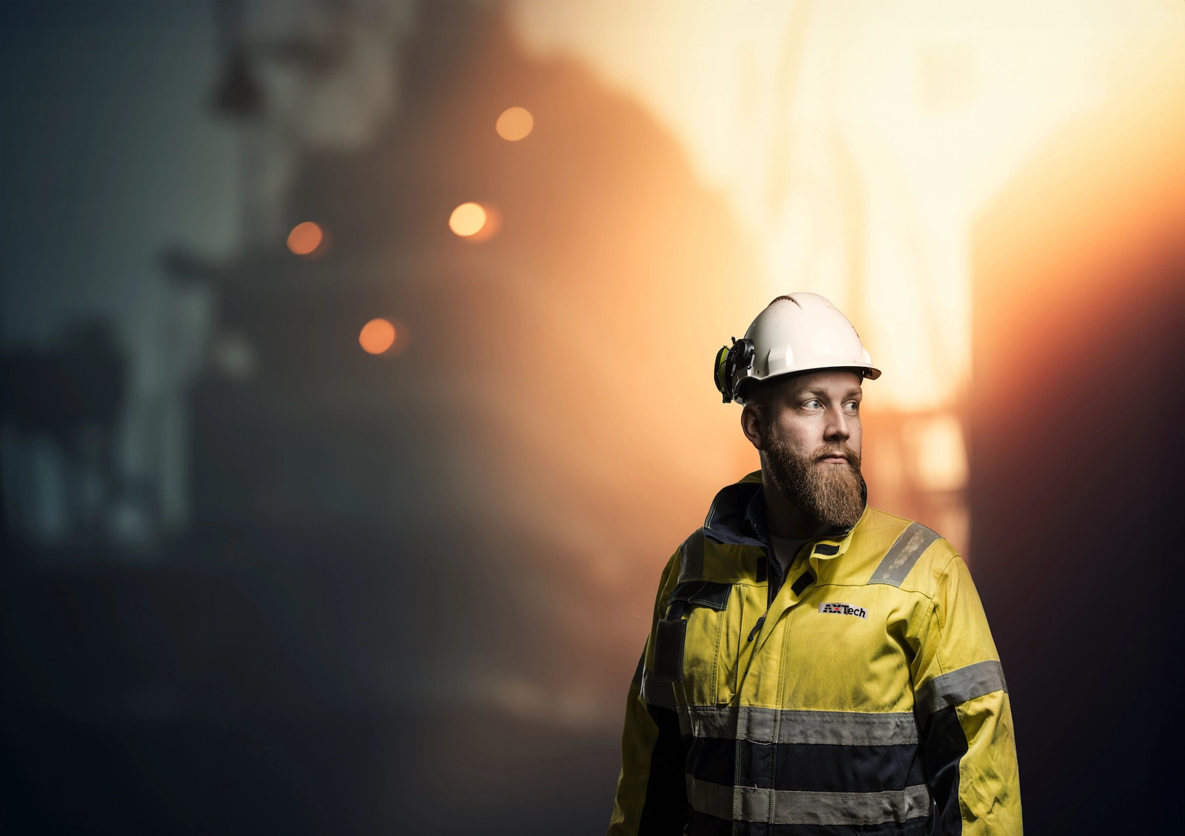 Bearded construction worker wearing a white hard hat and yellow safety jacket looking to the side at a dimly lit industrial site.