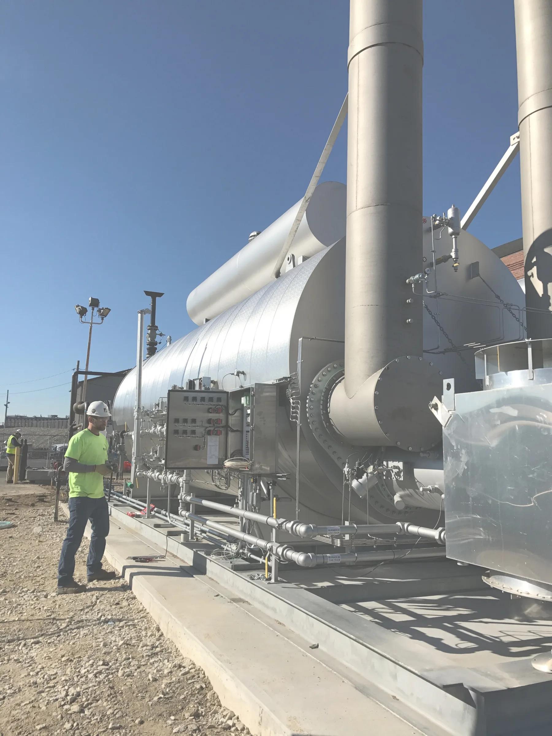 A technician in a hard hat stands at the burner control panels of a large gas-fired unit on a new concrete pad