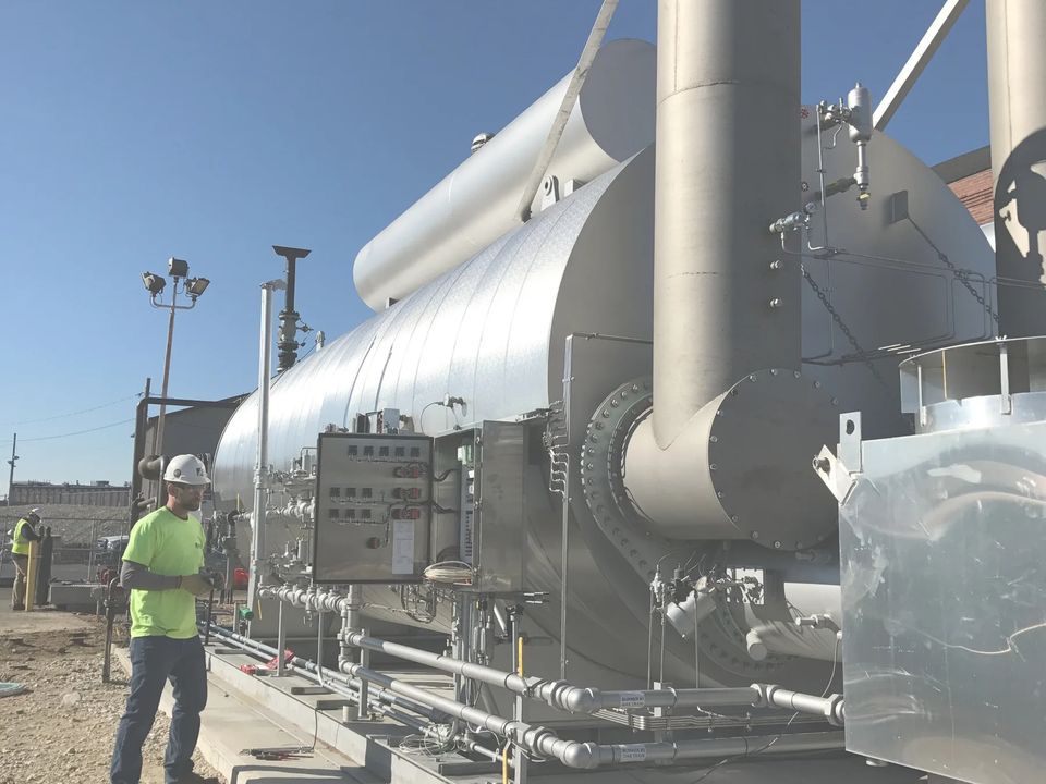A technician in a hard hat stands at the burner control panels of a large gas-fired unit on a new concrete pad