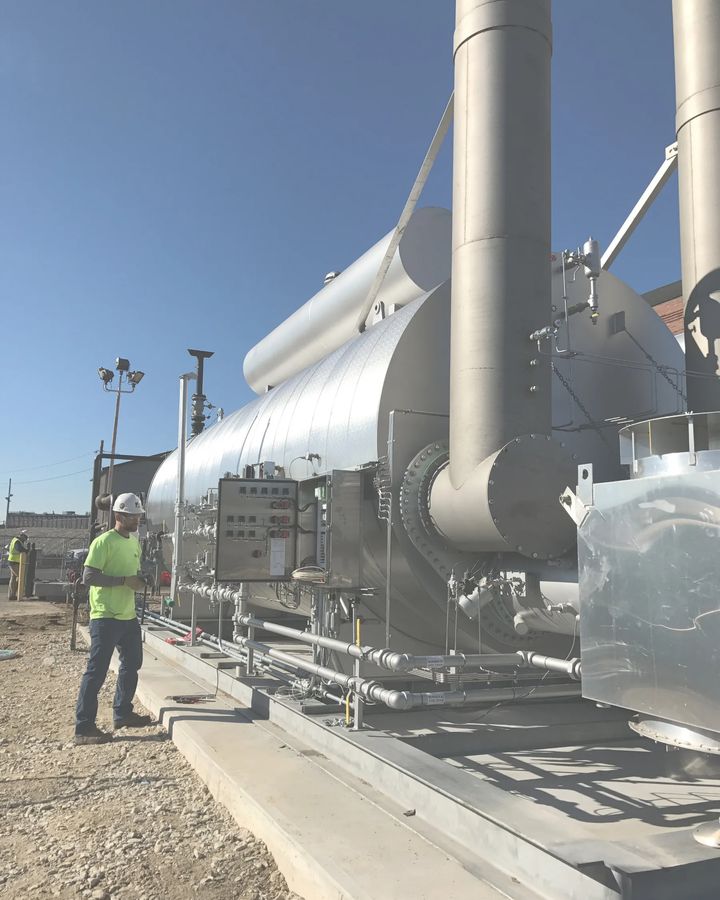 A technician in a hard hat stands at the burner control panels of a large gas-fired unit on a new concrete pad