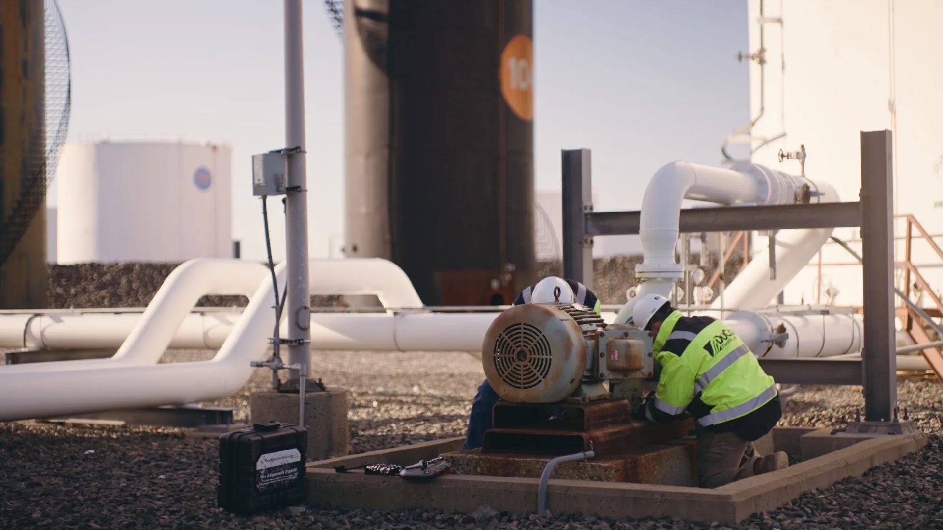 A technician in hi-vis kneels beside a pump motor between white pipes and storage tanks