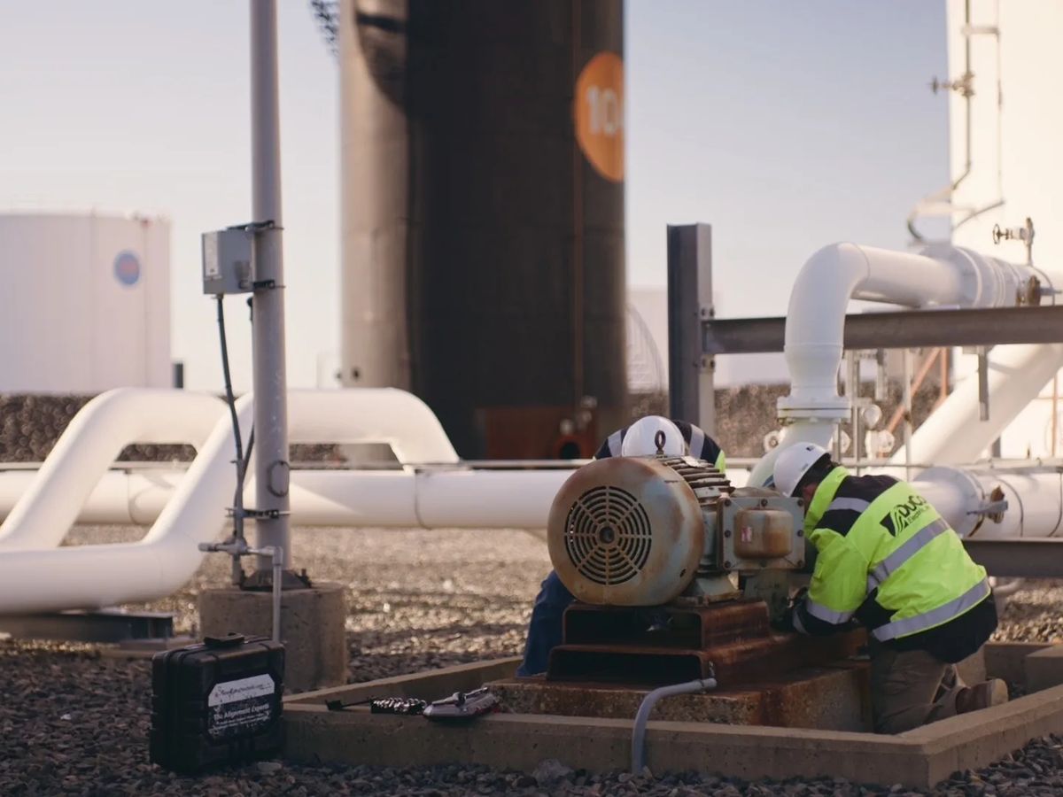 A technician in hi-vis kneels beside a pump motor between white pipes and storage tanks