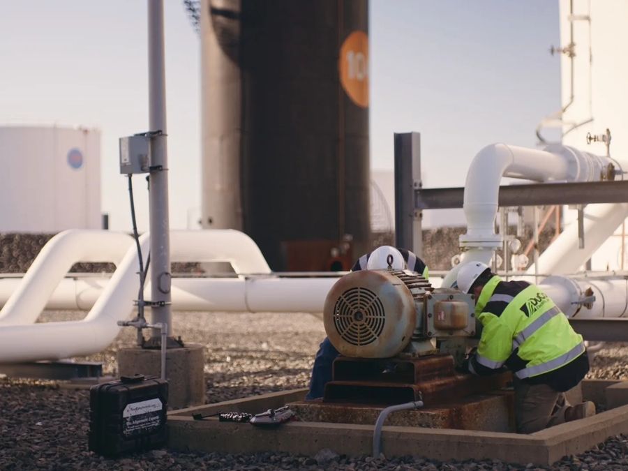 A technician in hi-vis kneels beside a pump motor between white pipes and storage tanks