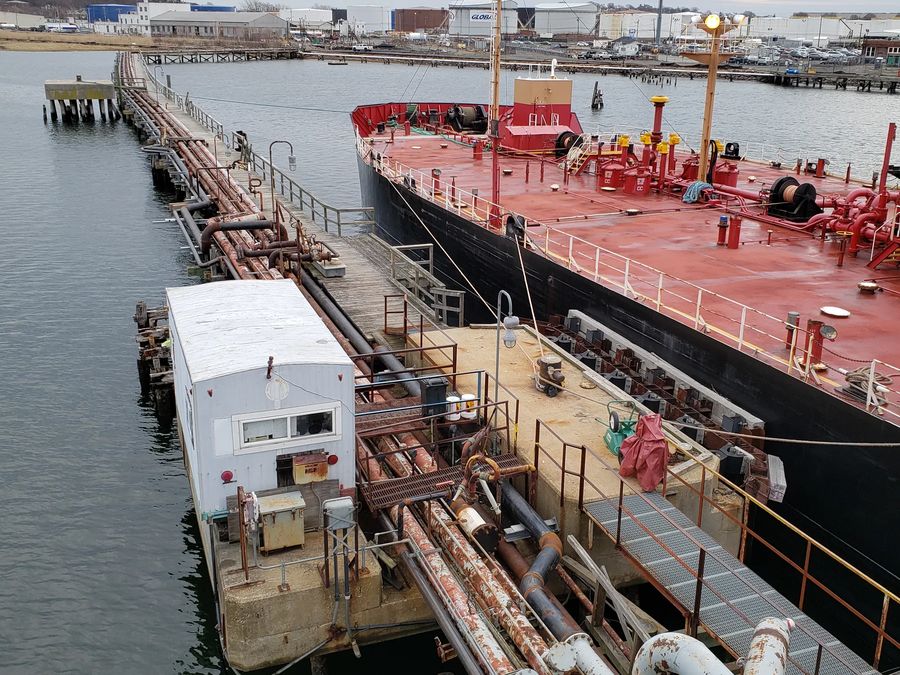 A tanker moored alongside a fuel pier, with pipelines and a small white building running along the dock