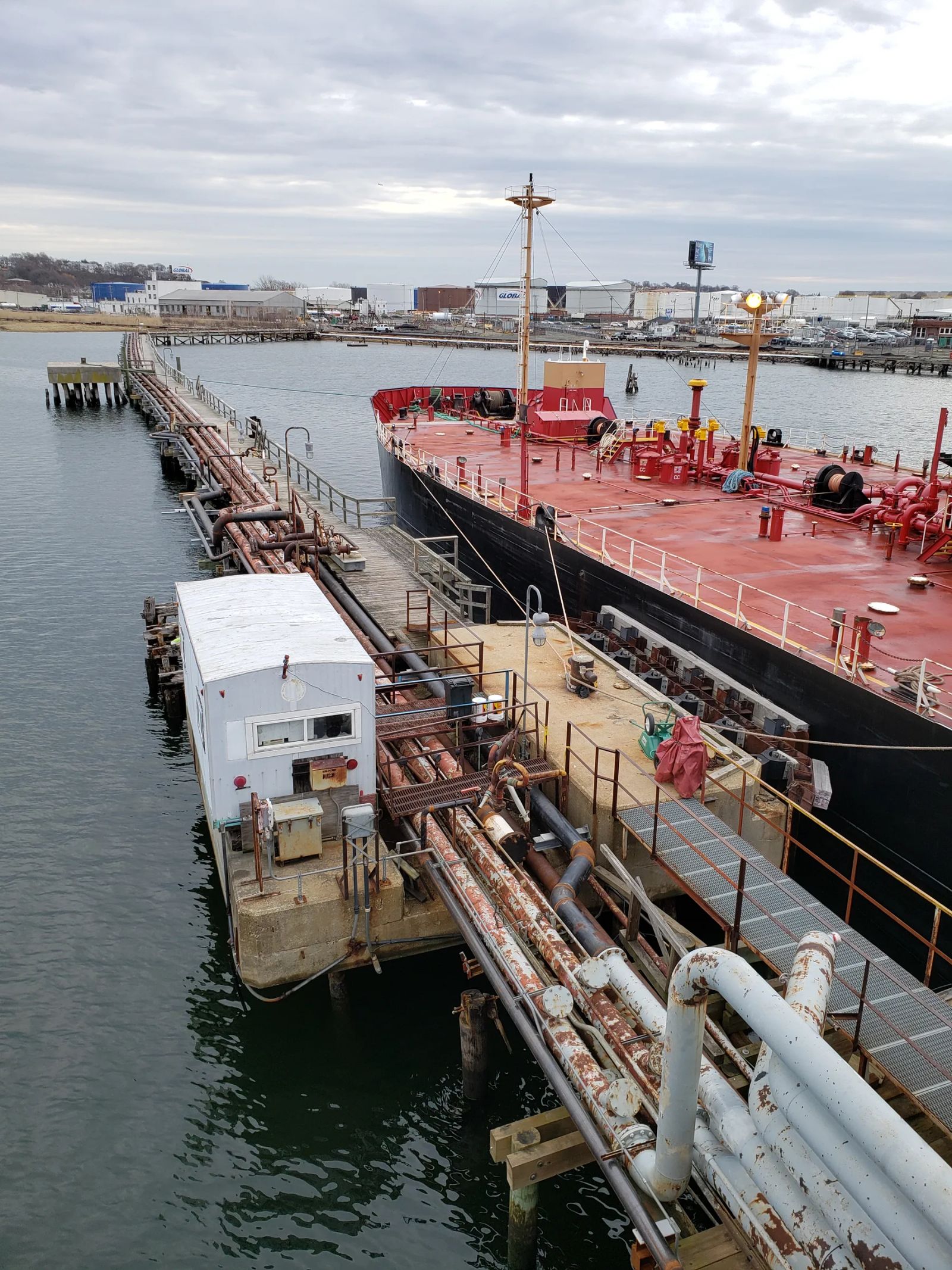 A tanker moored alongside a fuel pier, with pipelines and a small white building running along the dock