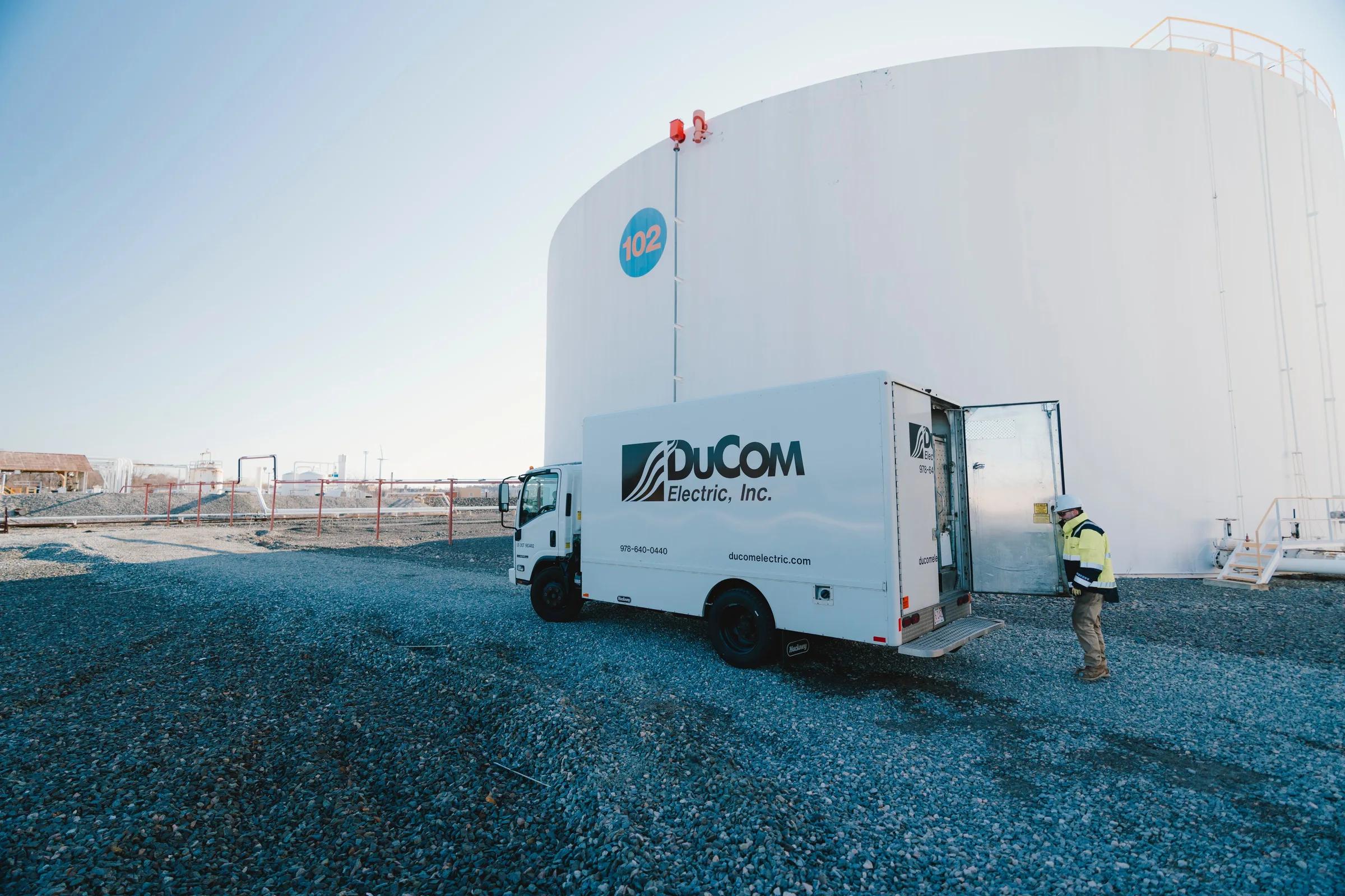 A DuCom Electric box truck parked on gravel beside a large white storage tank, with a technician in hi-vis at the open rear door